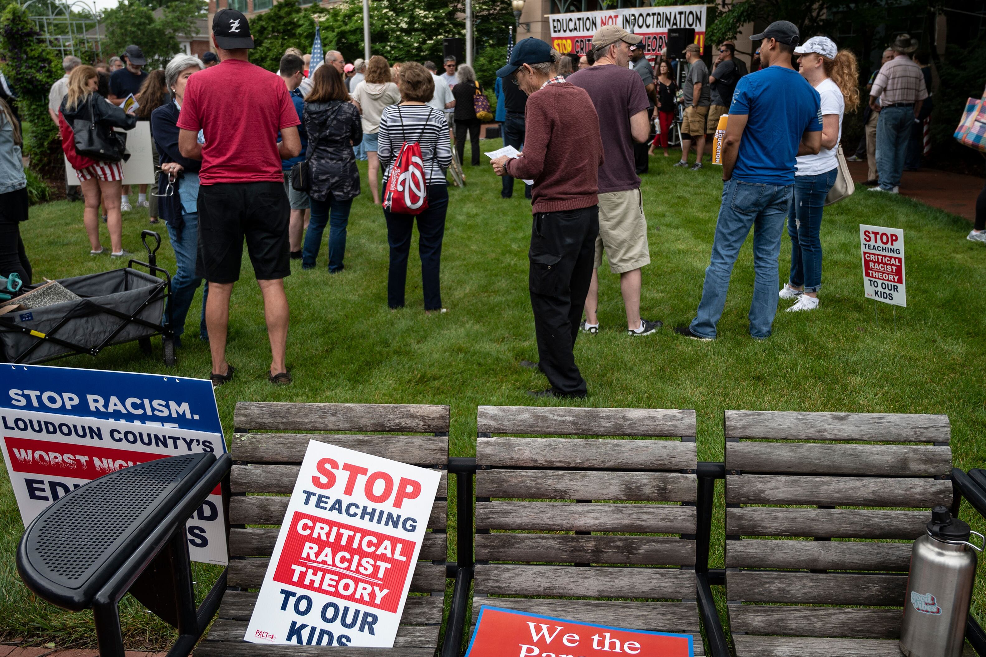 A group of people stand in the grass while a set of wooden chairs holds signs that read "stop teaching critical race theory to our kids."