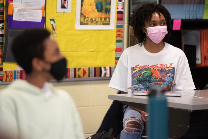 Students sit in a Chicago classroom, both with a mask on and a yellow bulletin board behind them. One student is in the foreground but blurry, the student in the background is sharper in the image.