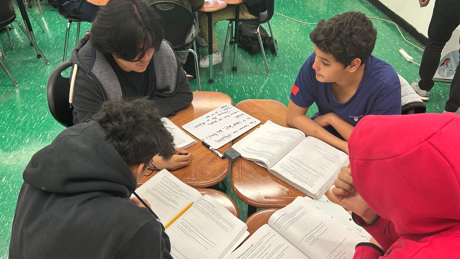 Four boys sit in a circle studying, with open schoolbooks in front of them. One wears a black hoodie and one wears a red hoodie over his head