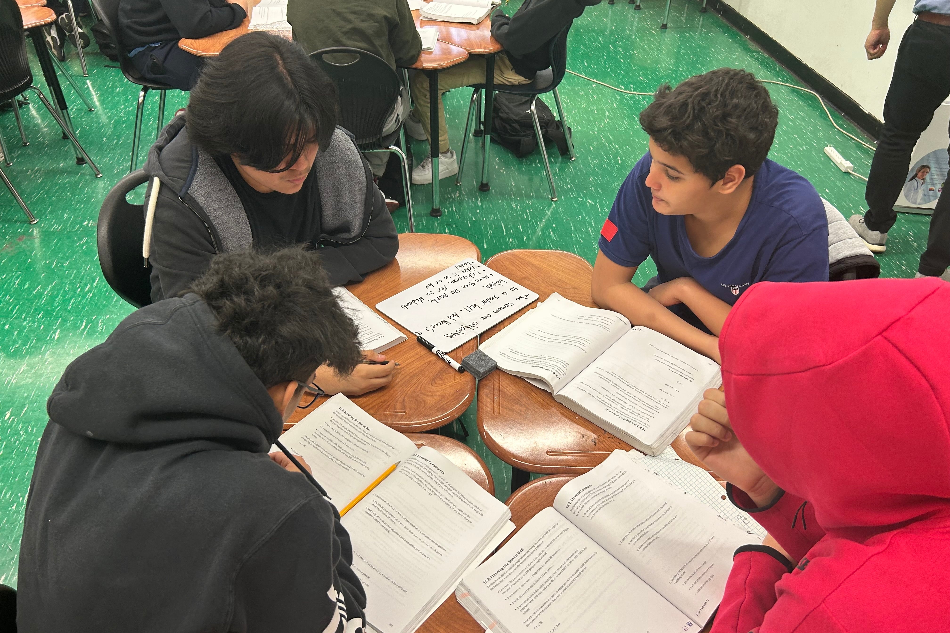 Four boys sit in a circle studying, with open schoolbooks in front of them. One wears a black hoodie and one wears a red hoodie over his head
