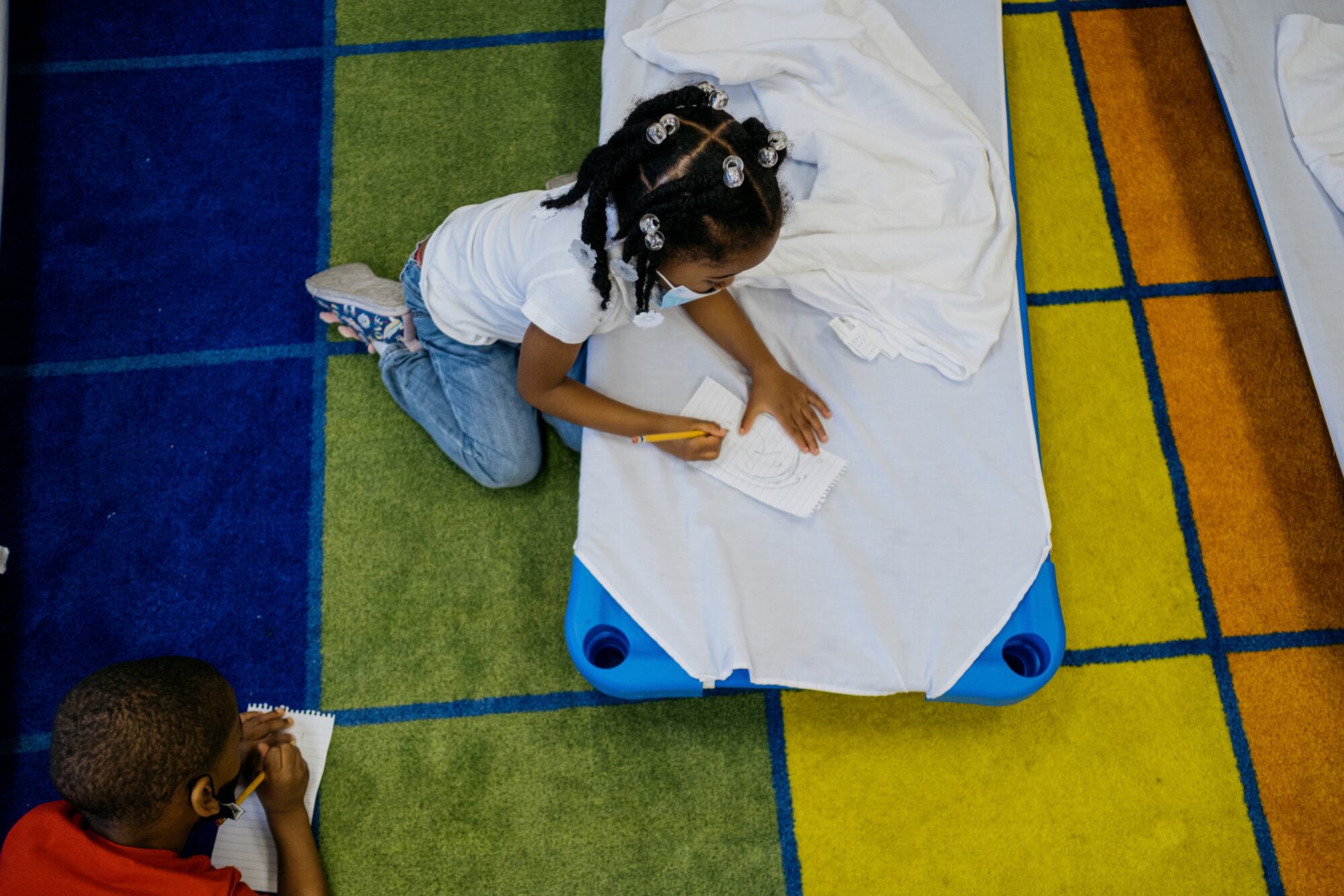 Two young students, seen from above, write on a piece of paper with a pencil while sitting on a colorful rug.