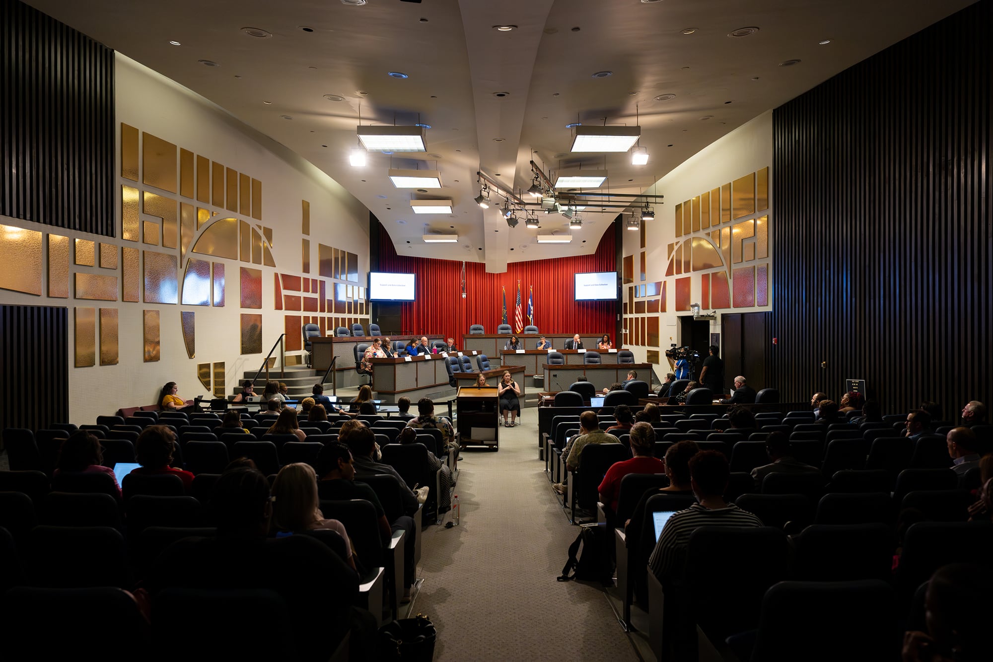 A photograph of a large meeting room with people sitting in rows of chairs facing a stage of people sitting in chairs.
