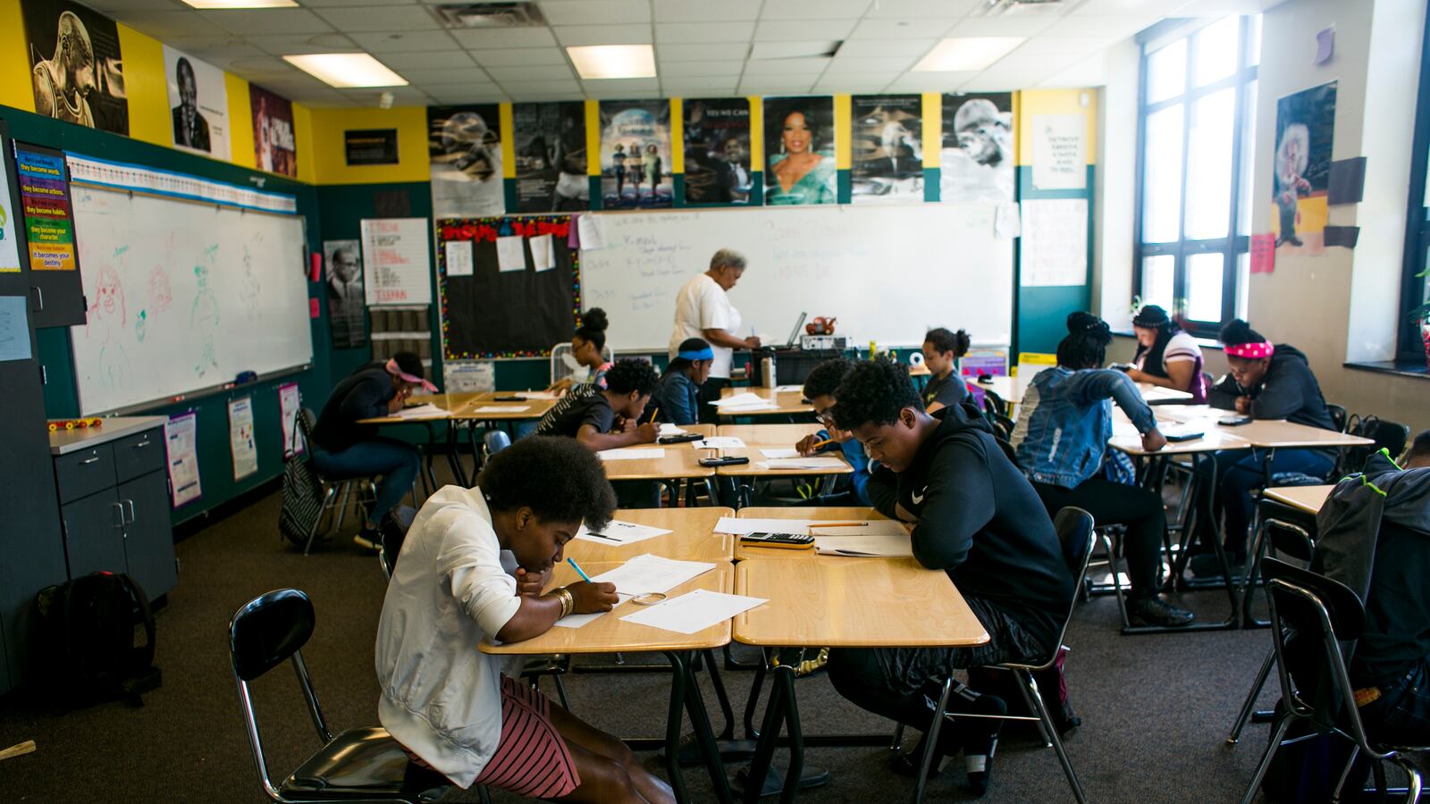 Students work at their their desks, which are grouped together, in a classroom as their teacher types on a computer at the front of the room.