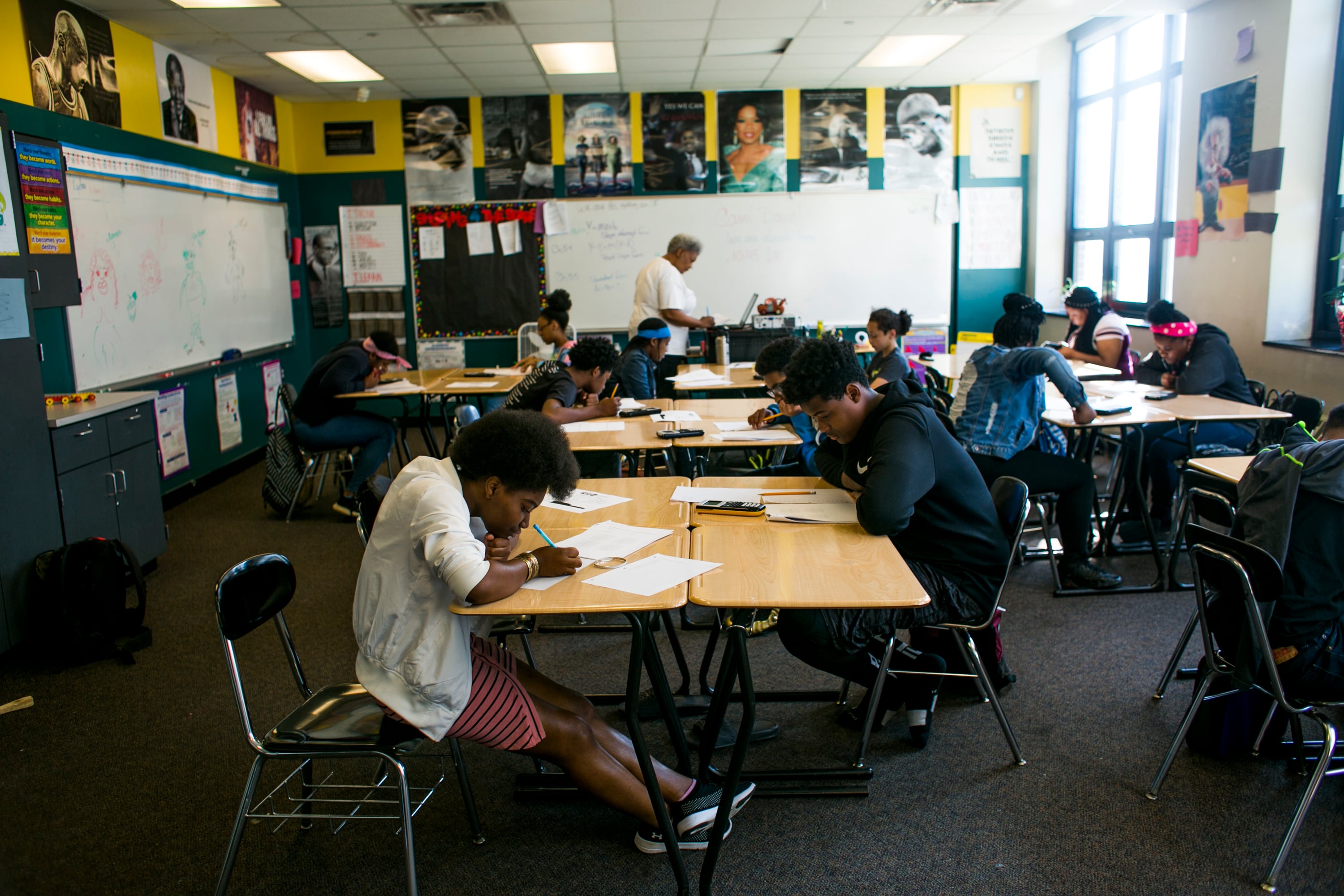 Students work at their their desks, which are grouped together, in a classroom as their teacher types on a computer at the front of the room.
