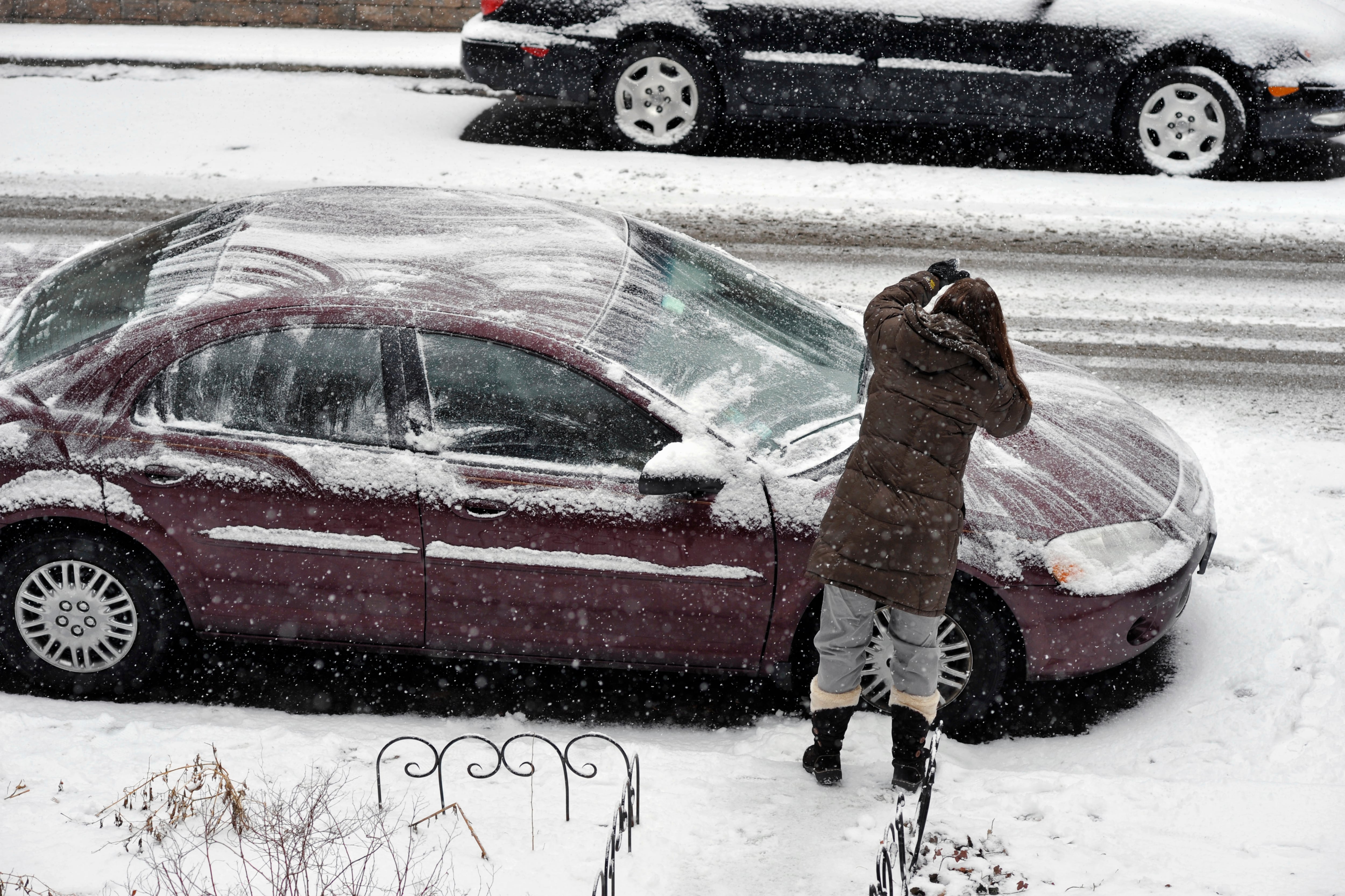 A woman in a dark coat cleans off the hood of a maroon car.