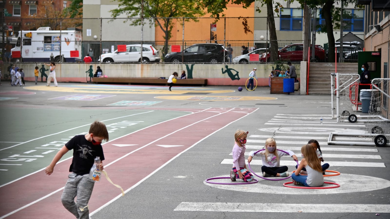 A young boy wearing a black shirt and grey pants walks along a track. Four girls play sitting within hula-hoops and many other children play in the background on a track during an after school program.