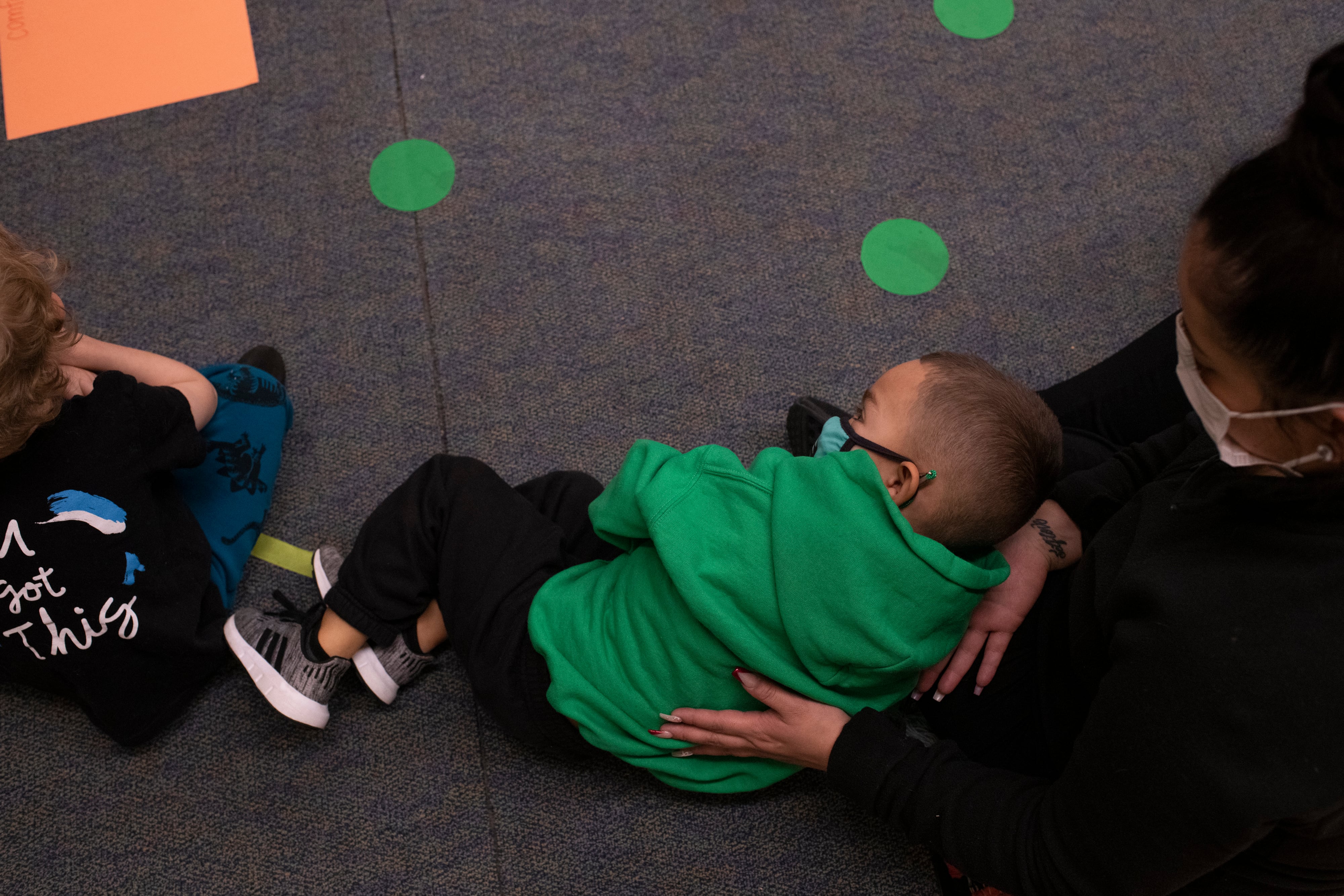 A preschool-aged boy leans against a paraprofessional while laying on the floor, each wearing masks, while the paraprofessional rests her hand on the boy’s back.