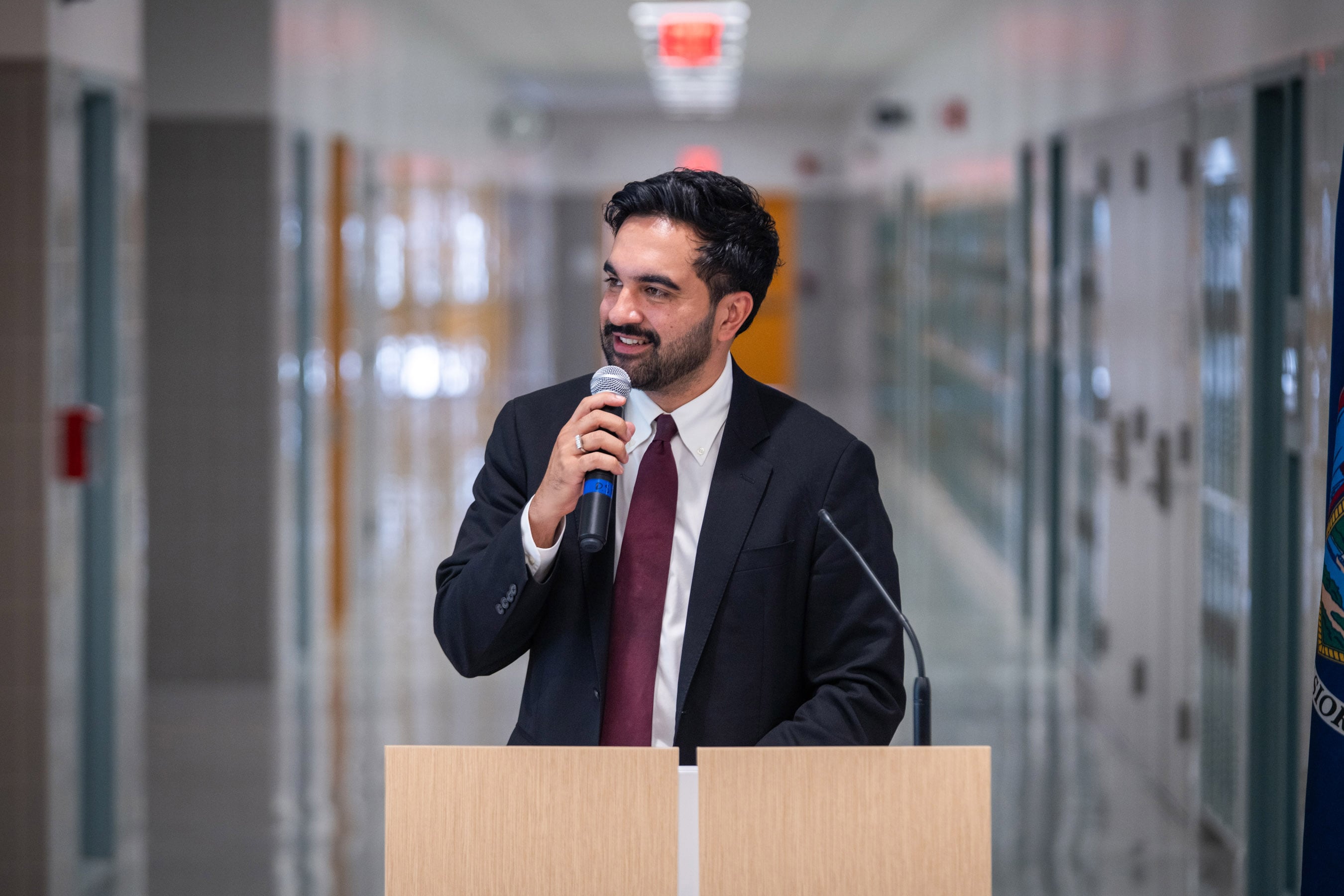 A man in a dark jacket and burgundy tie speaks into a microphone in a school hallway.