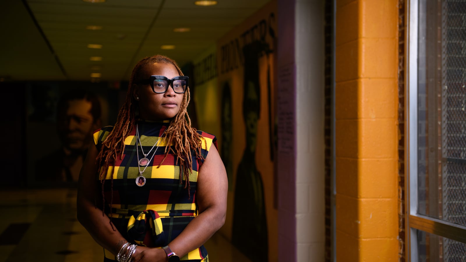 A Black woman in a striped dress looks out a window from a dark hallway inside a school.