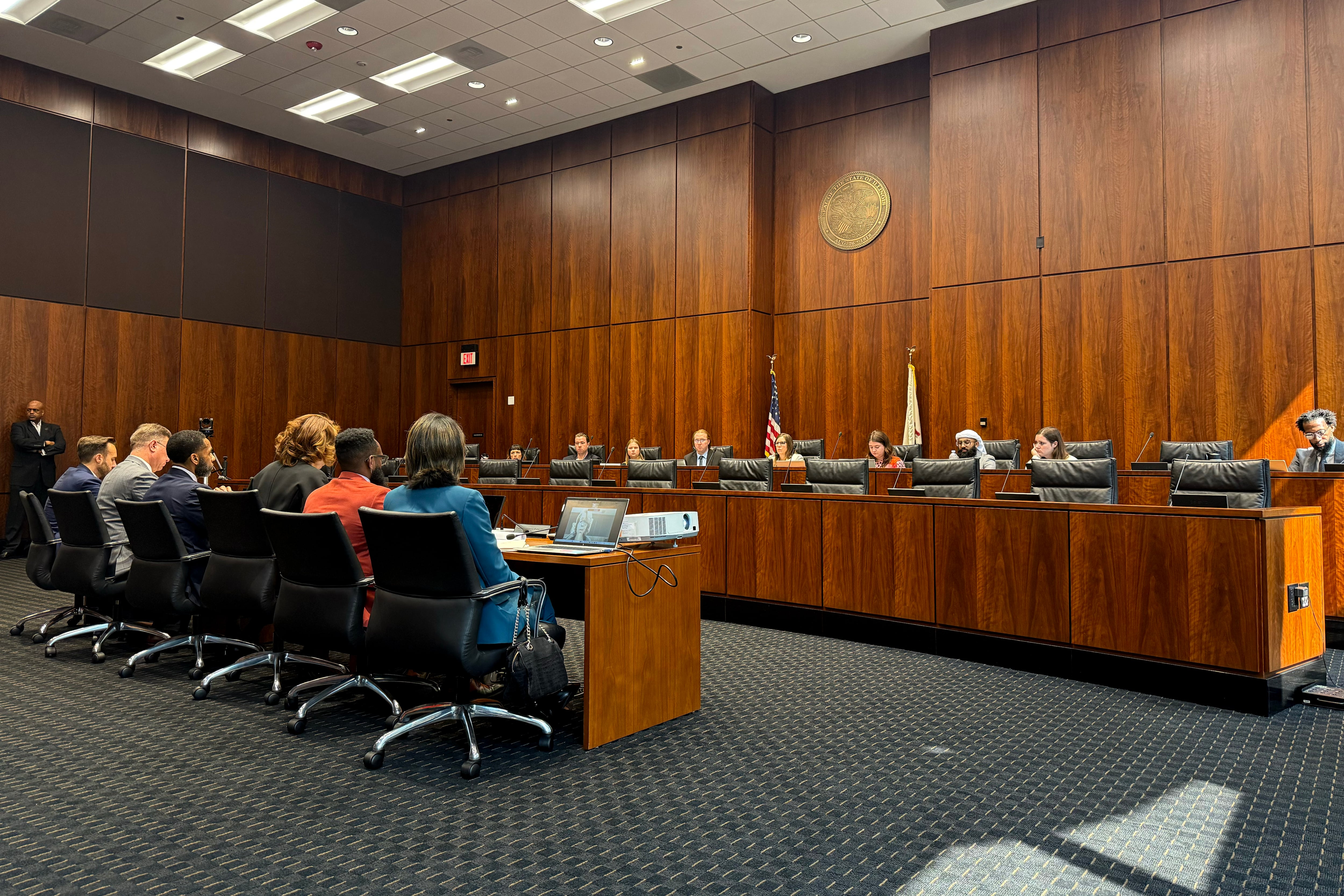 A photograph of two rows of wooden seats with people sitting in the two rows in a large conference room.