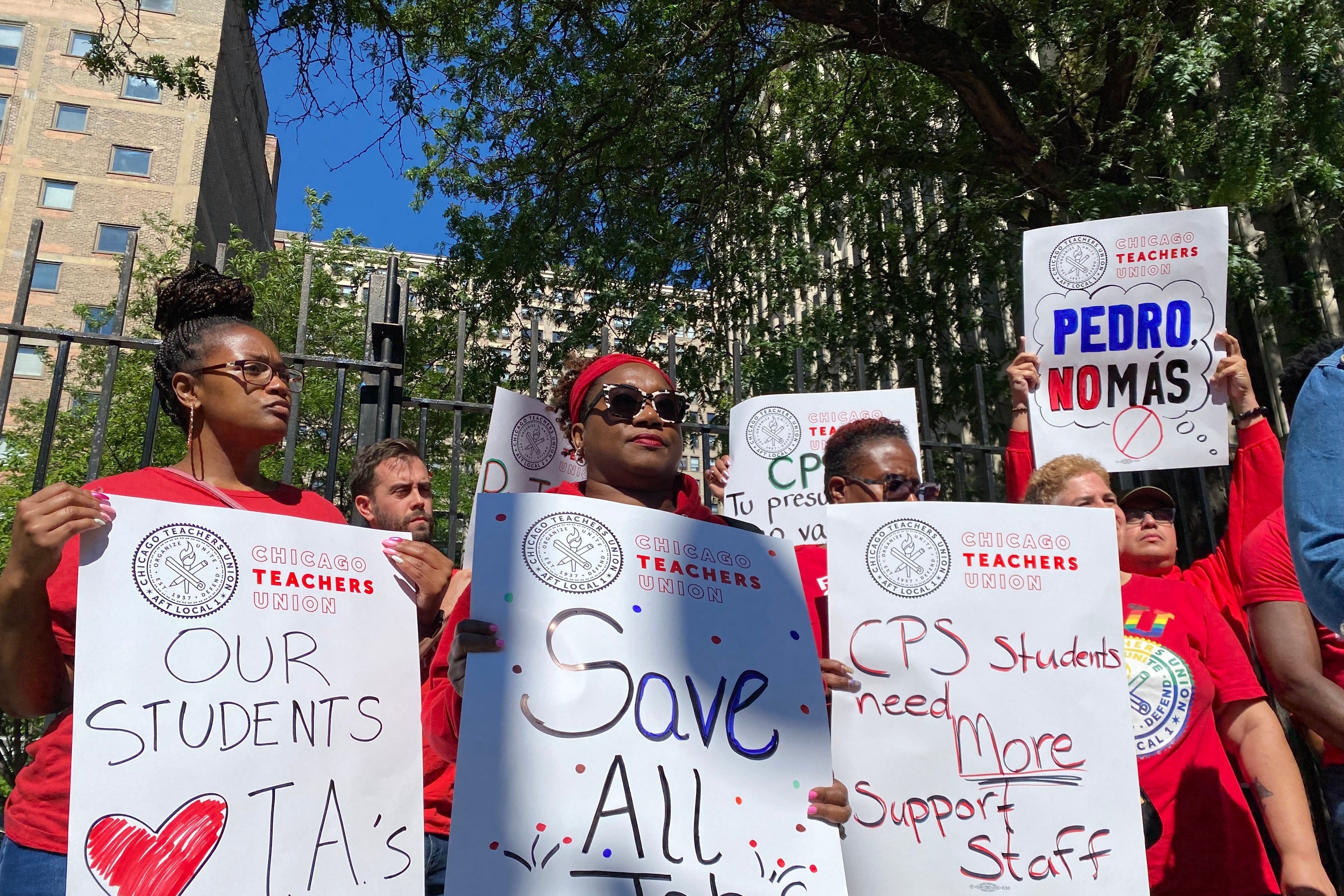 A group of adults wearing red t-shirts and holding protest signs stand outside.