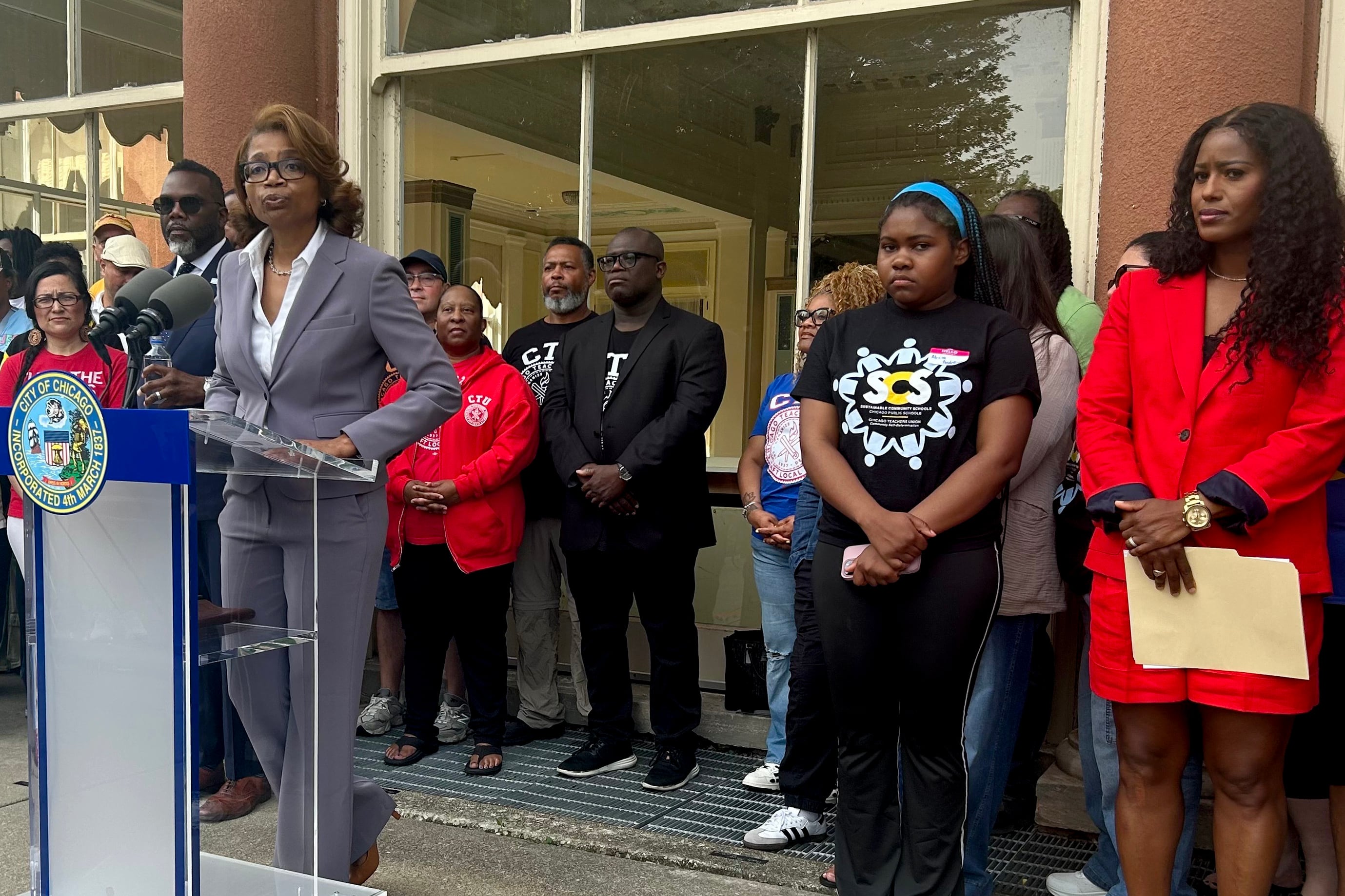 A group of adults stand outside of a large building while one person speaks from a podium.