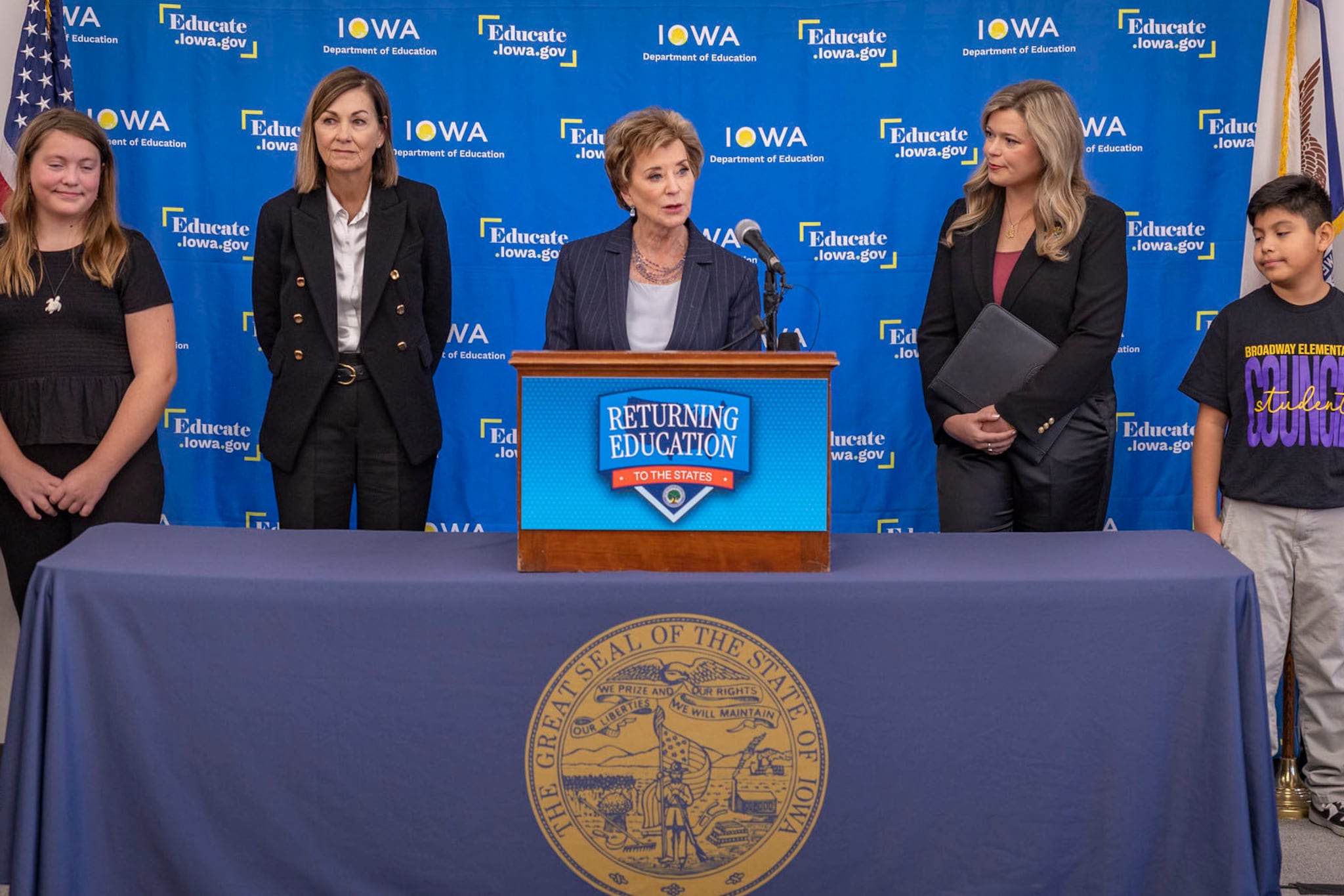 A photograph of a group of children standing on stage next to adults who are speaking from behind a podium.