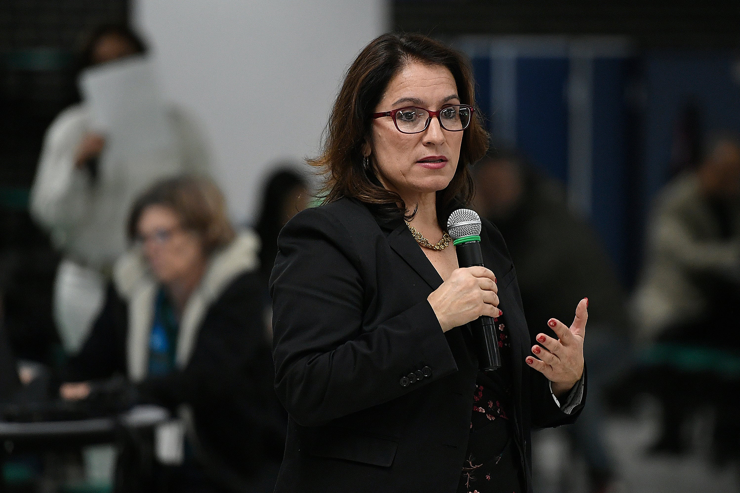 A woman with dark hair and wearing glasses holds a microphone with a crowd of people sitting and standing in the background.