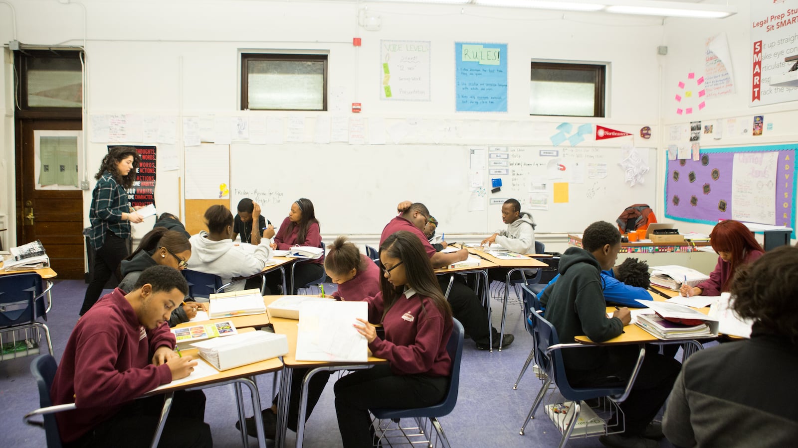 Students work at their desks in a classroom, all wearing maroon school uniforms.