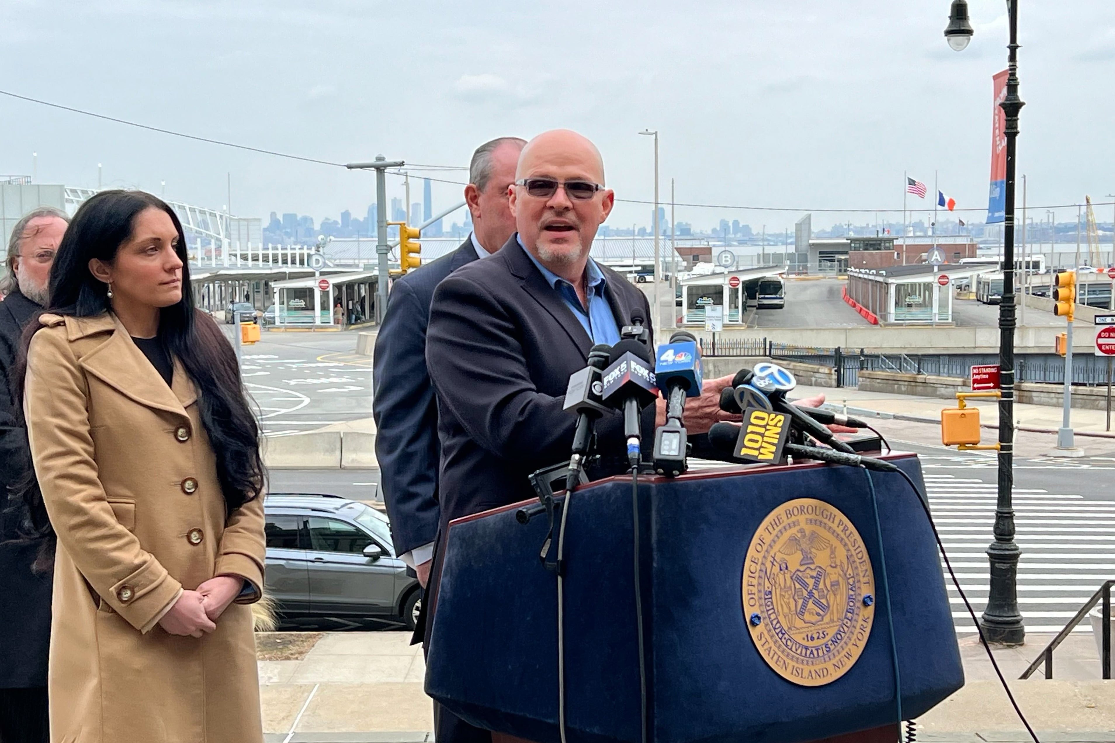 A man wearing a dark suit stands at a podium with three people standing behind him and a street scene and cityscape in the background.