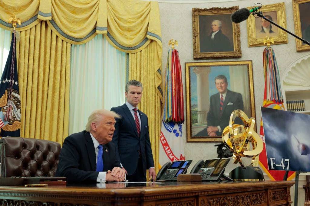 President Donald Trump, an older man in a dark suit with a blue tie, sits at an ornate desk in a lavish office. It's the Oval Office of the White House. Another man in a suit stands next to him. Behind them are heavy gold curtains and the portraits of former presidents.