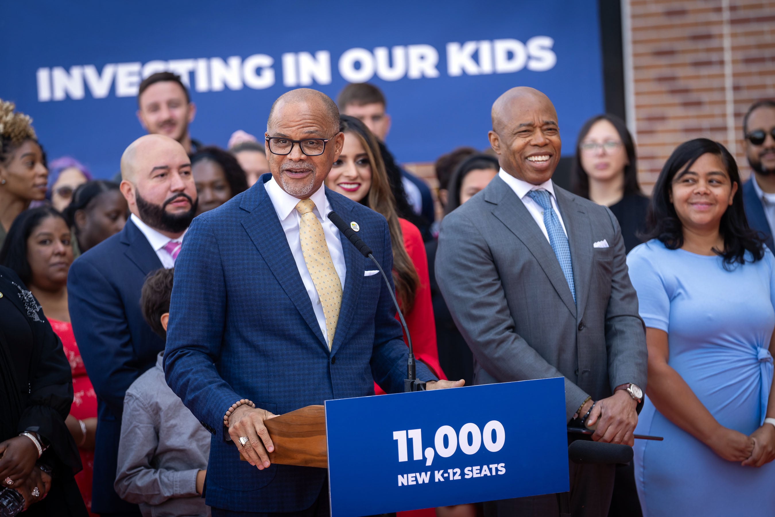 People in suits and business clothes stand behind a wooden podium with a blue paper sign and in front of a crowd of people outside.