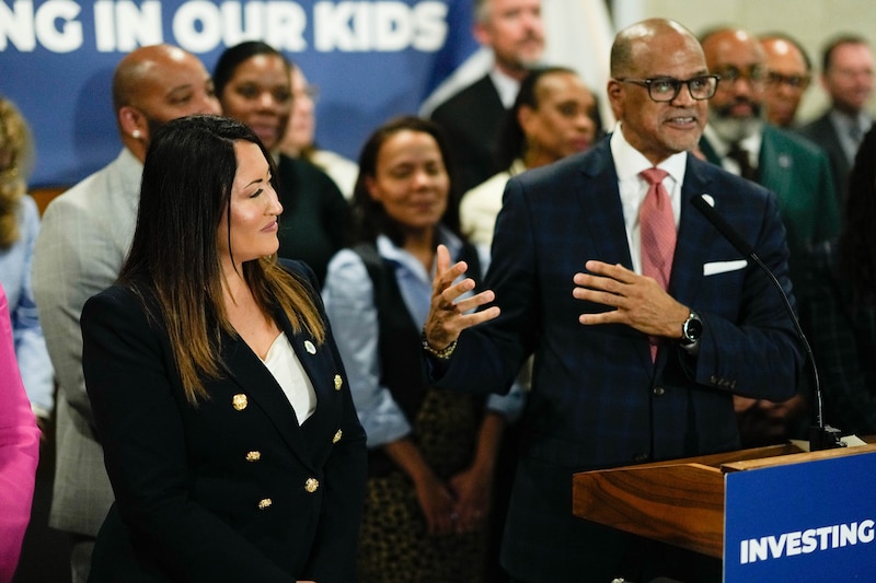 Two people in suits speak from behind a podium with a large group of people behind them.