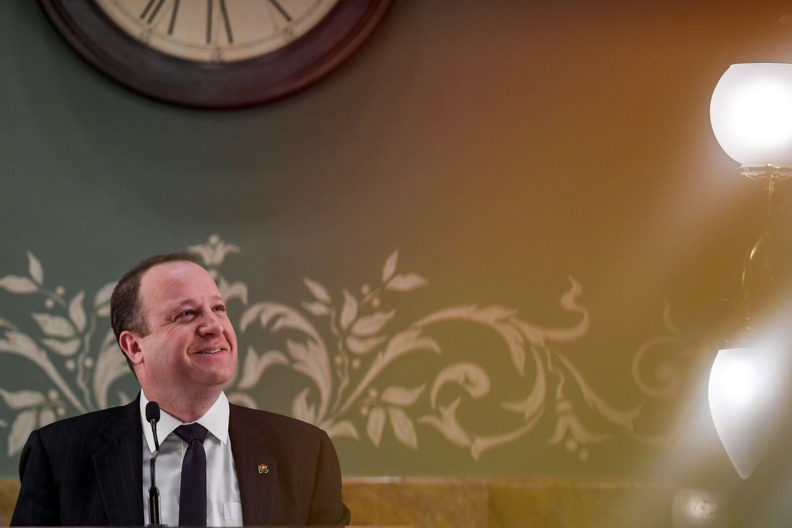 Governor Jared Polis stands at a podium in front of a green wall. Large bright lights glare to the right of the governor.