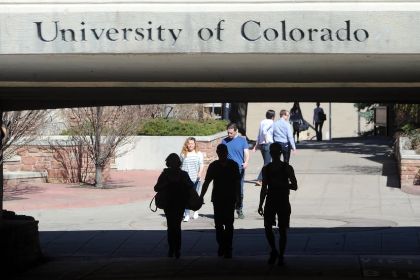 Several people walk underneath a grey bridge with the words “University of Colorado” etched into the concrete.