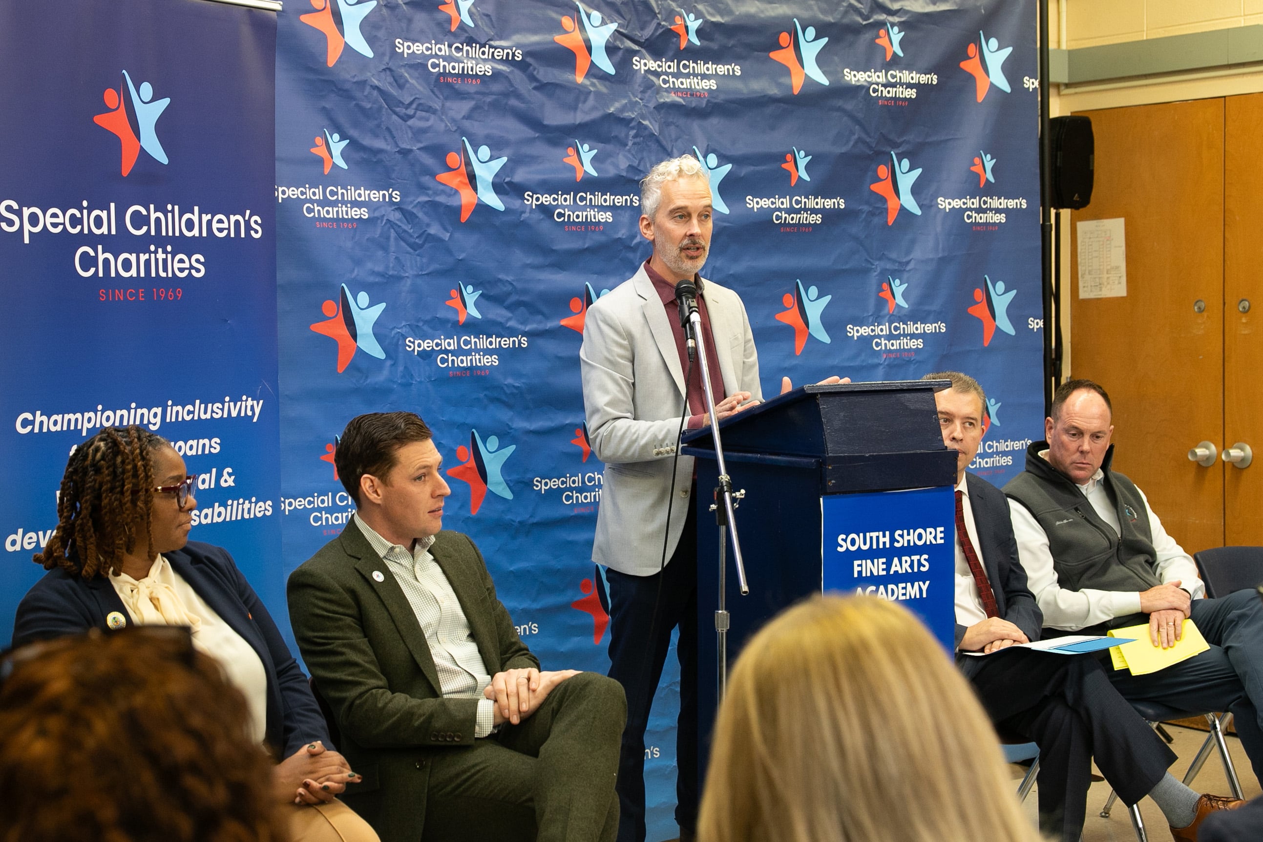 A man in a suit speaks from a podium in front of a colorful background and next to four adults sitting in chairs.