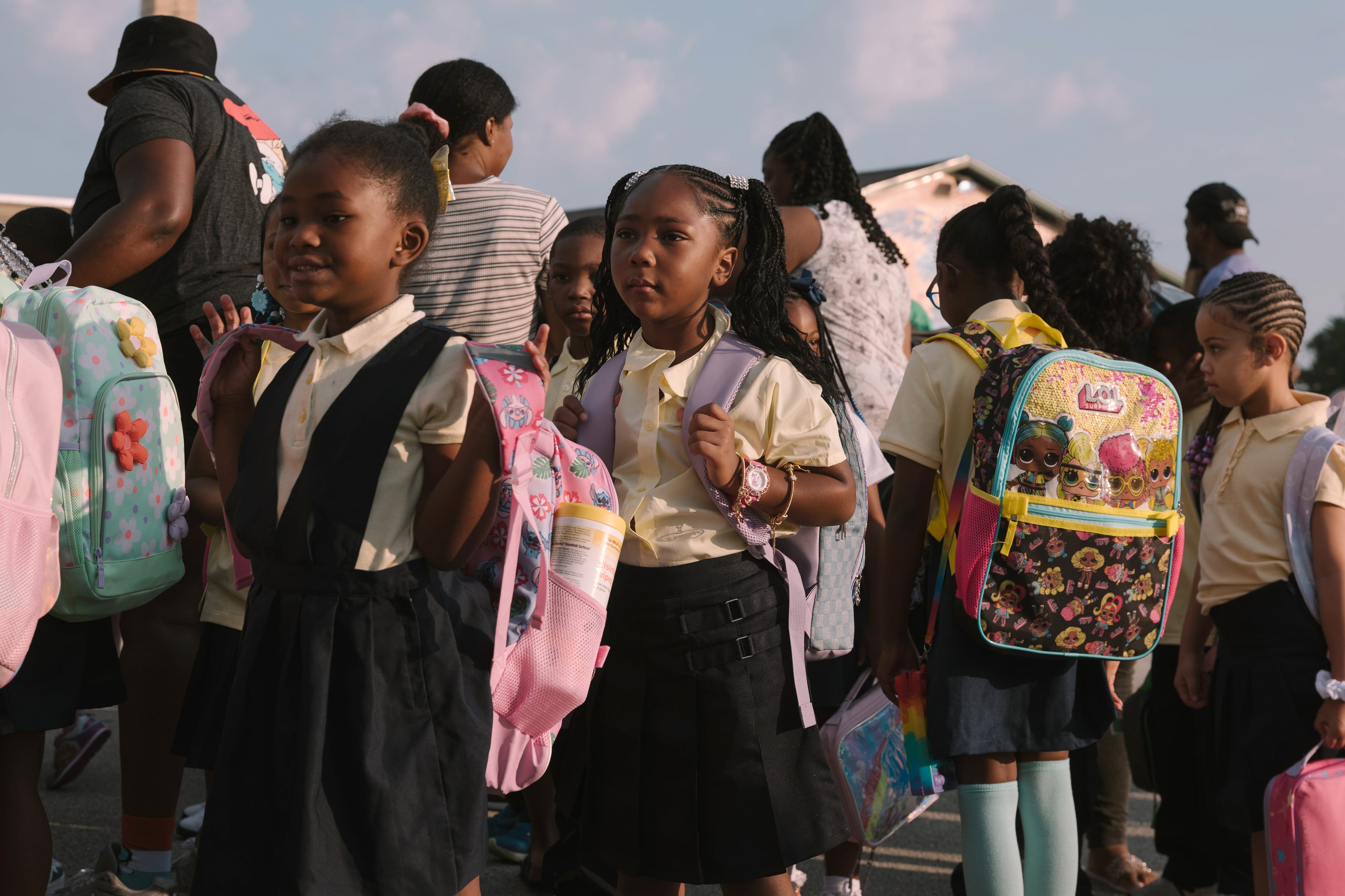 A row of young students stand outside with their backpacks with a clear sky in the background.
