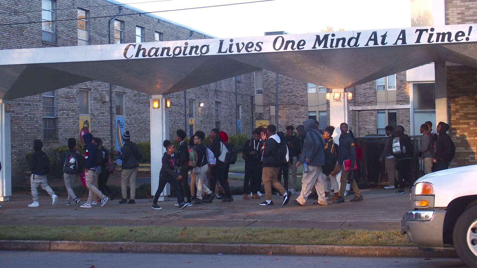 Students wearing backpacks walk under a school awning that says, “Changing lives one mind at a time!”