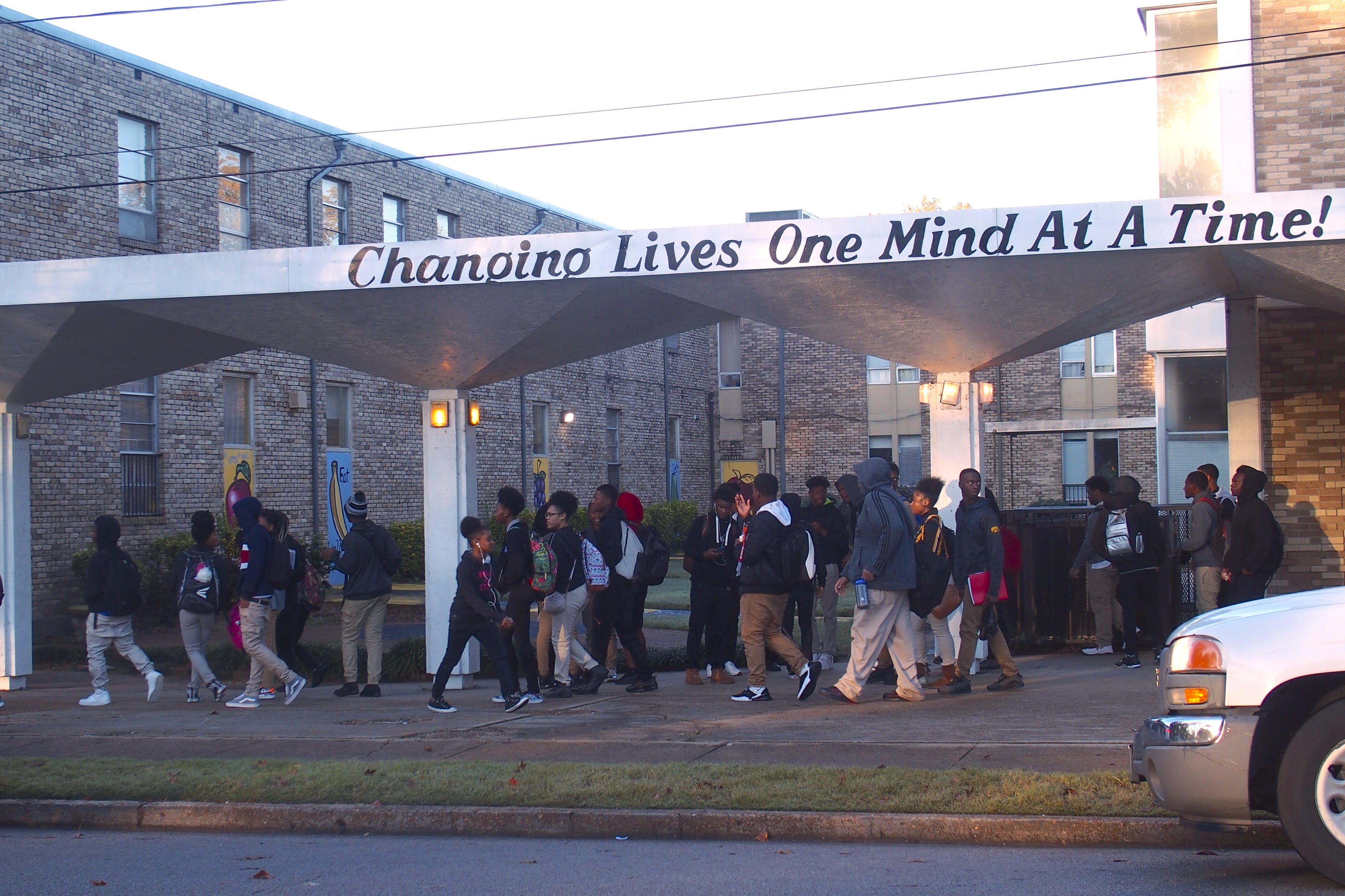 Students wearing backpacks walk under a school awning that says, “Changing lives one mind at a time!” 