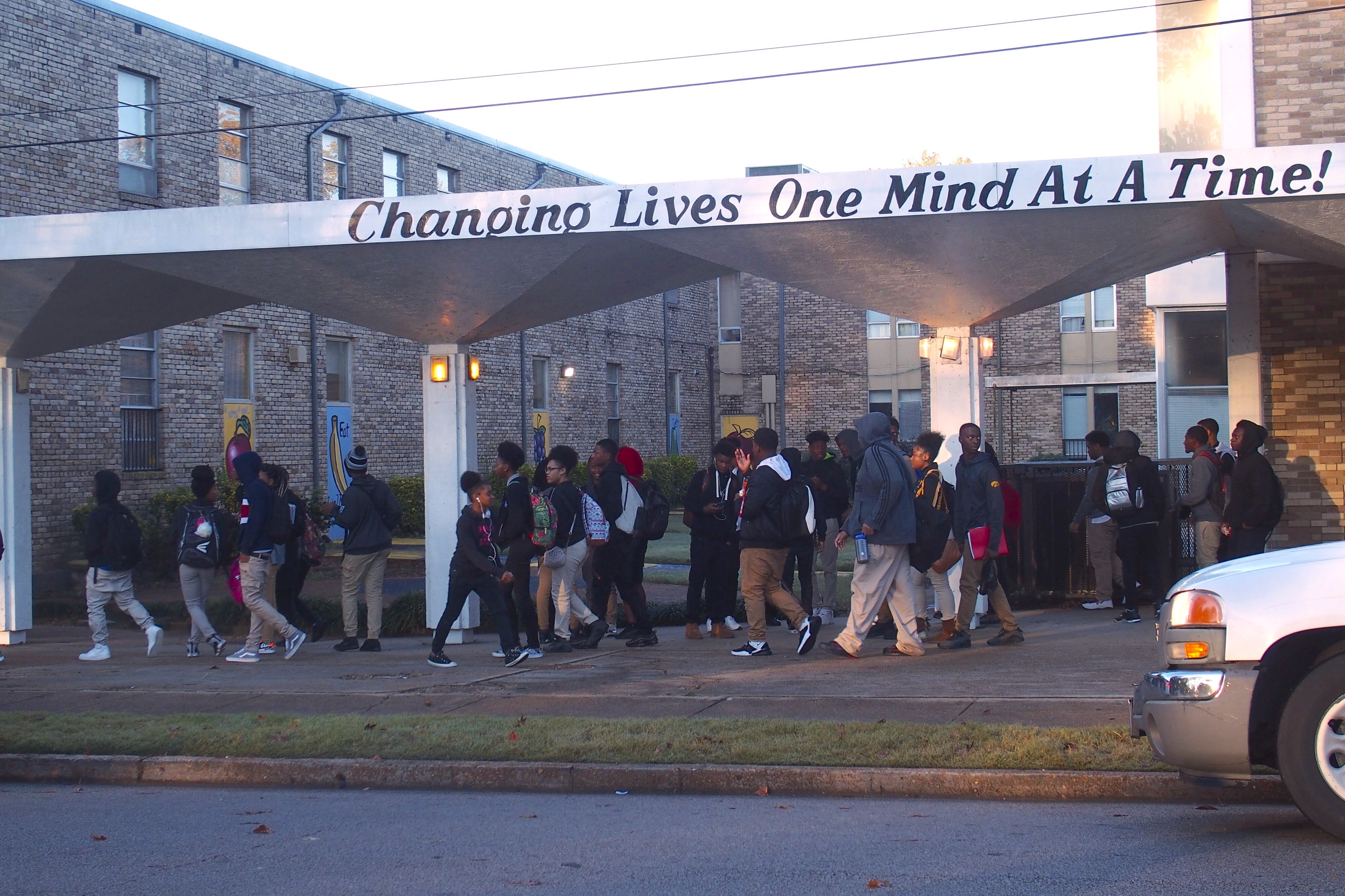 Students wearing backpacks walk under a school awning that says, “Changing lives one mind at a time!”
