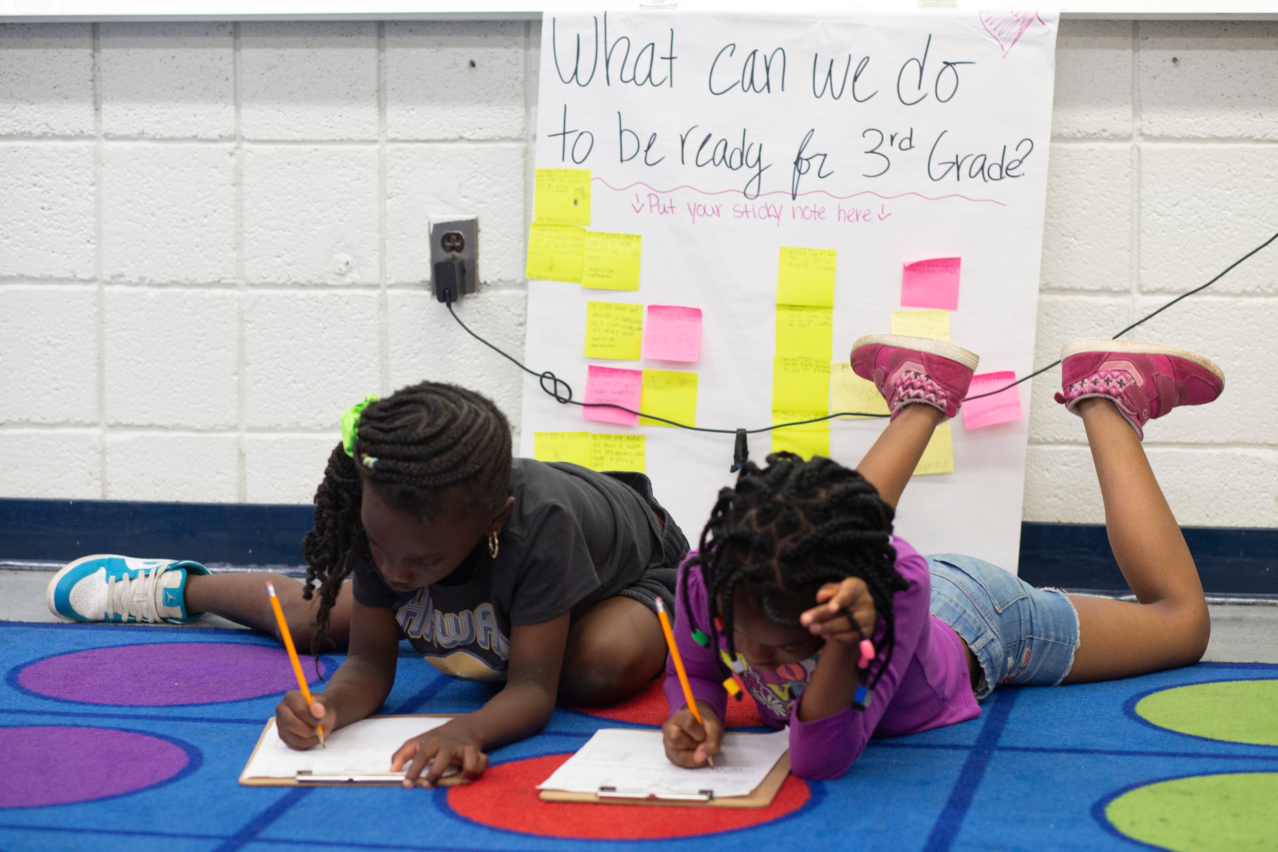 Two girls lay on a blue rug holding a pencil as they write on a piece of paper.