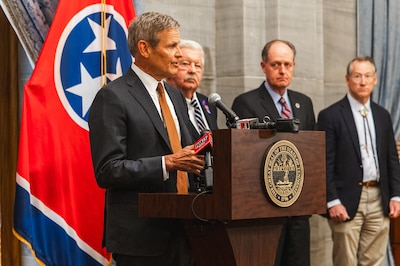 A man in a dark suit stands behind a wooden podium with a Tennessee state flag and three men standing in the background.