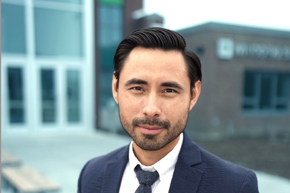 Headshot of a man with black hair and a beard. He is wearing a dark suit and tie and a white shirt. Buildings are in the background.