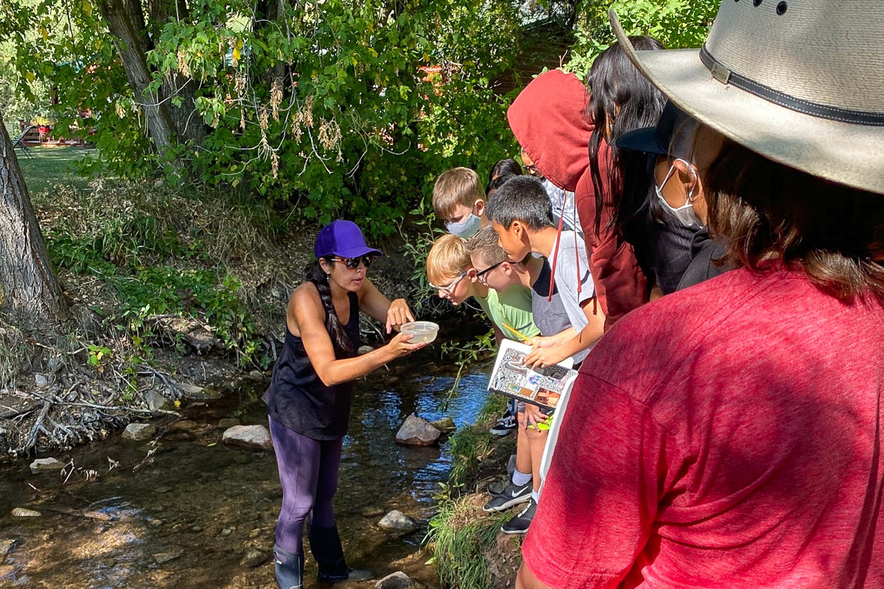 A teacher works with her students at a creek, with her children looking at something she is holding in a small container.