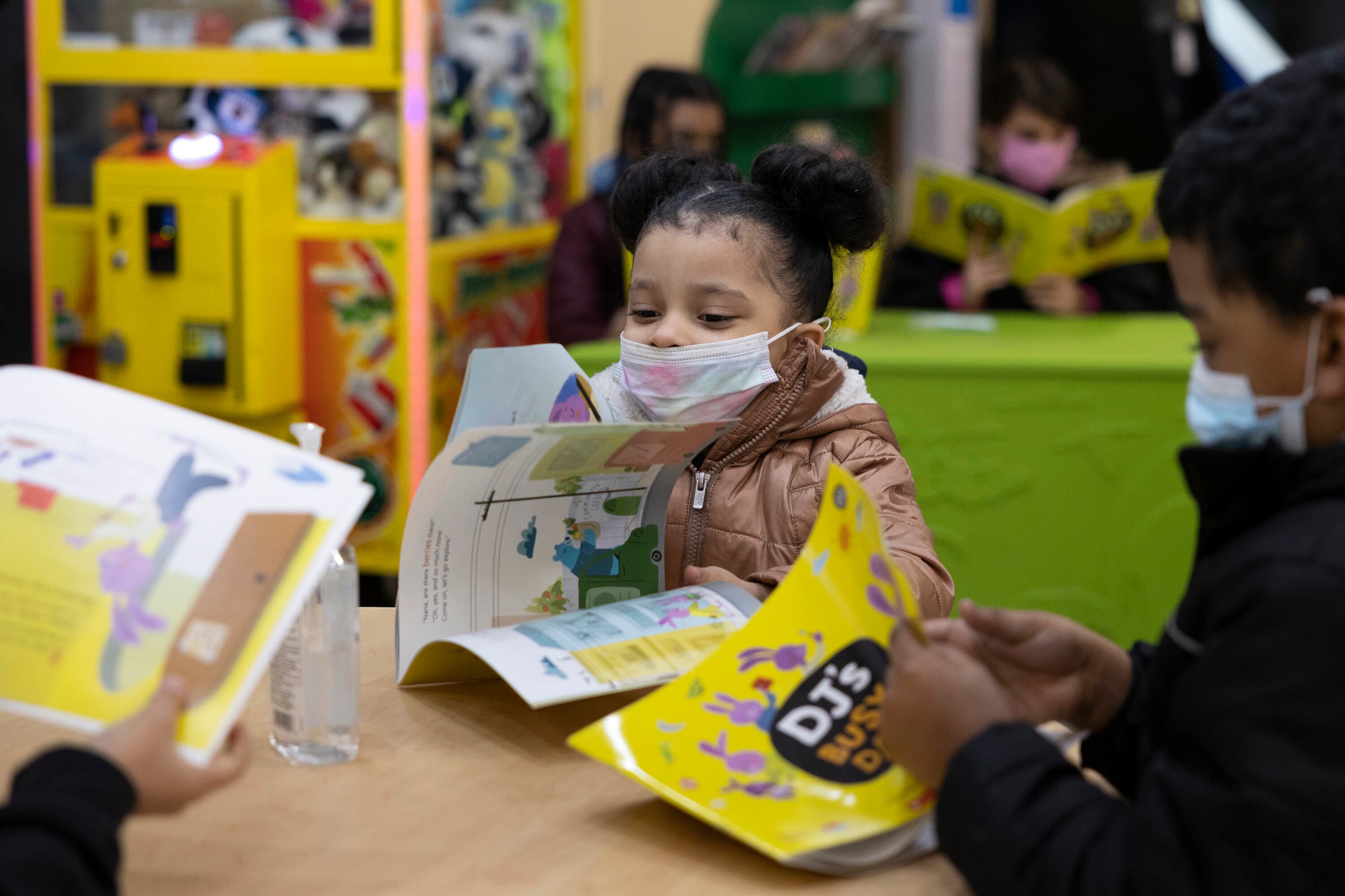 A little girl wearing a white and pink mask reads a children’s book at a desk while two others at the desk read “DJ’s Busy Day.”