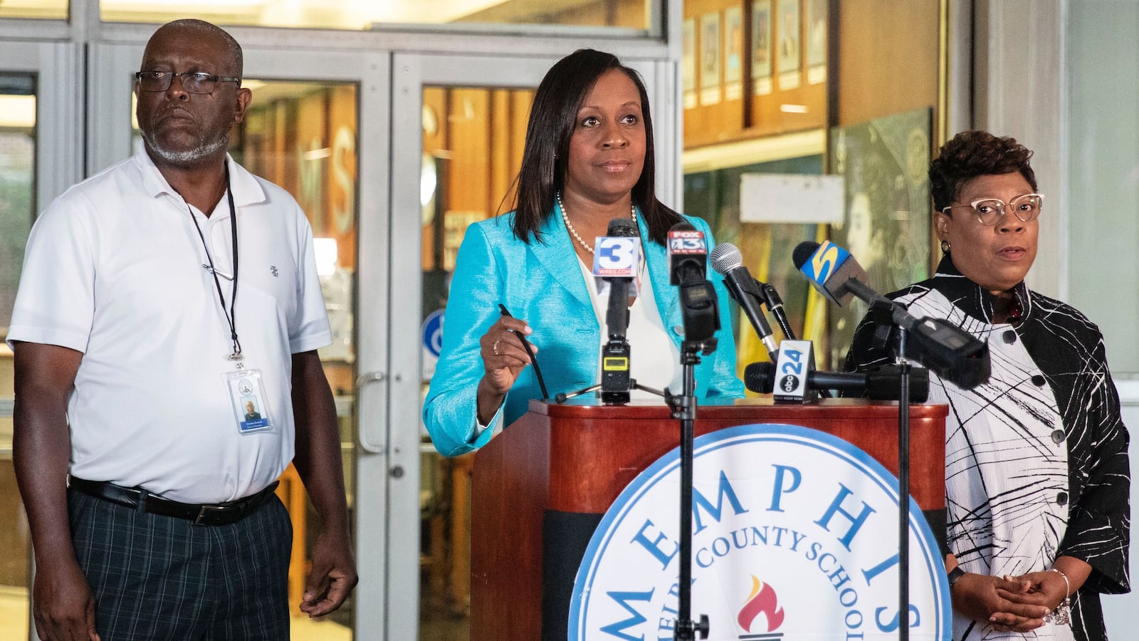 A woman stands in front of a podium with TV news microphones, with a man and woman standing beside her