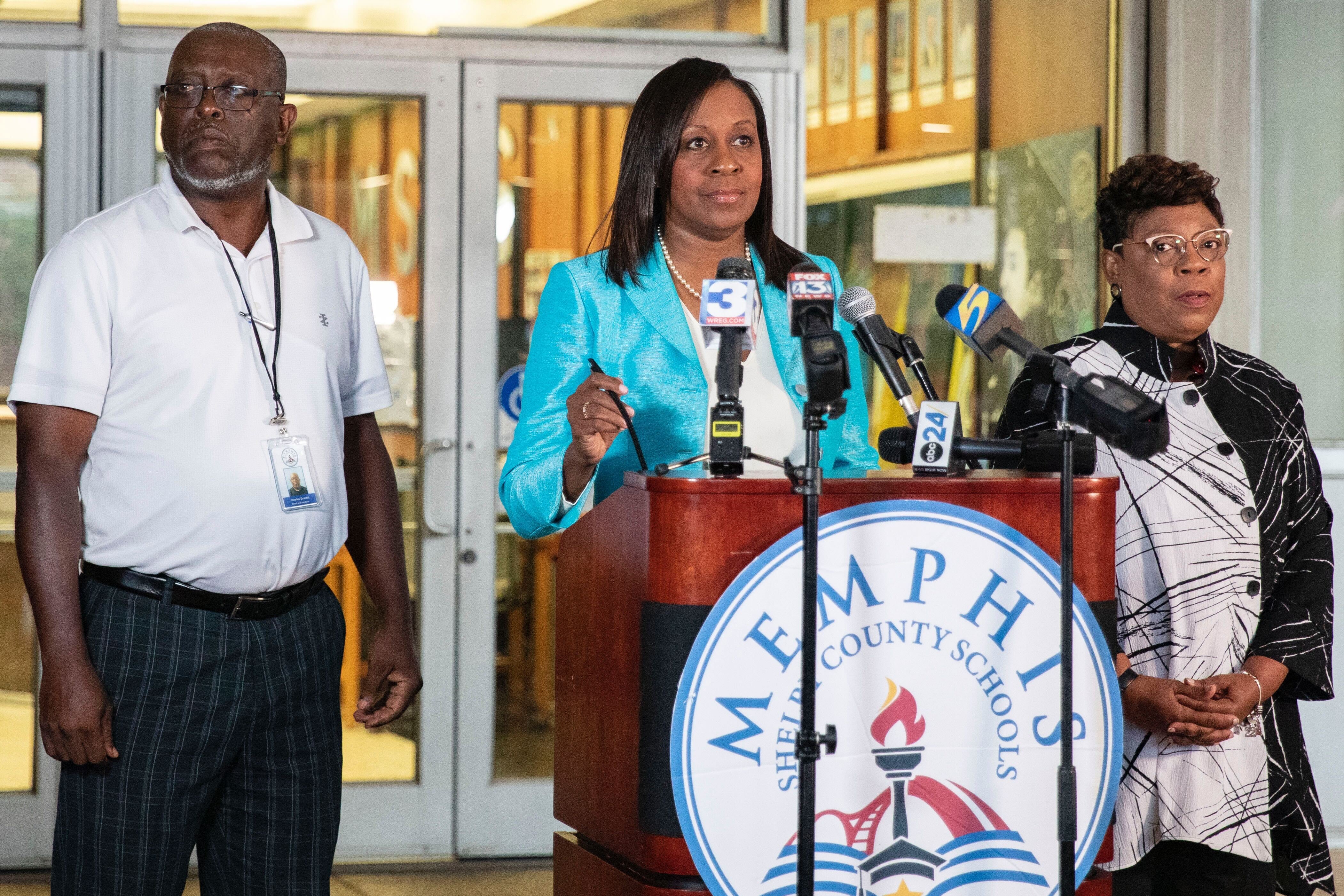 A woman stands in front of a podium with TV news microphones, with a man and woman standing beside her