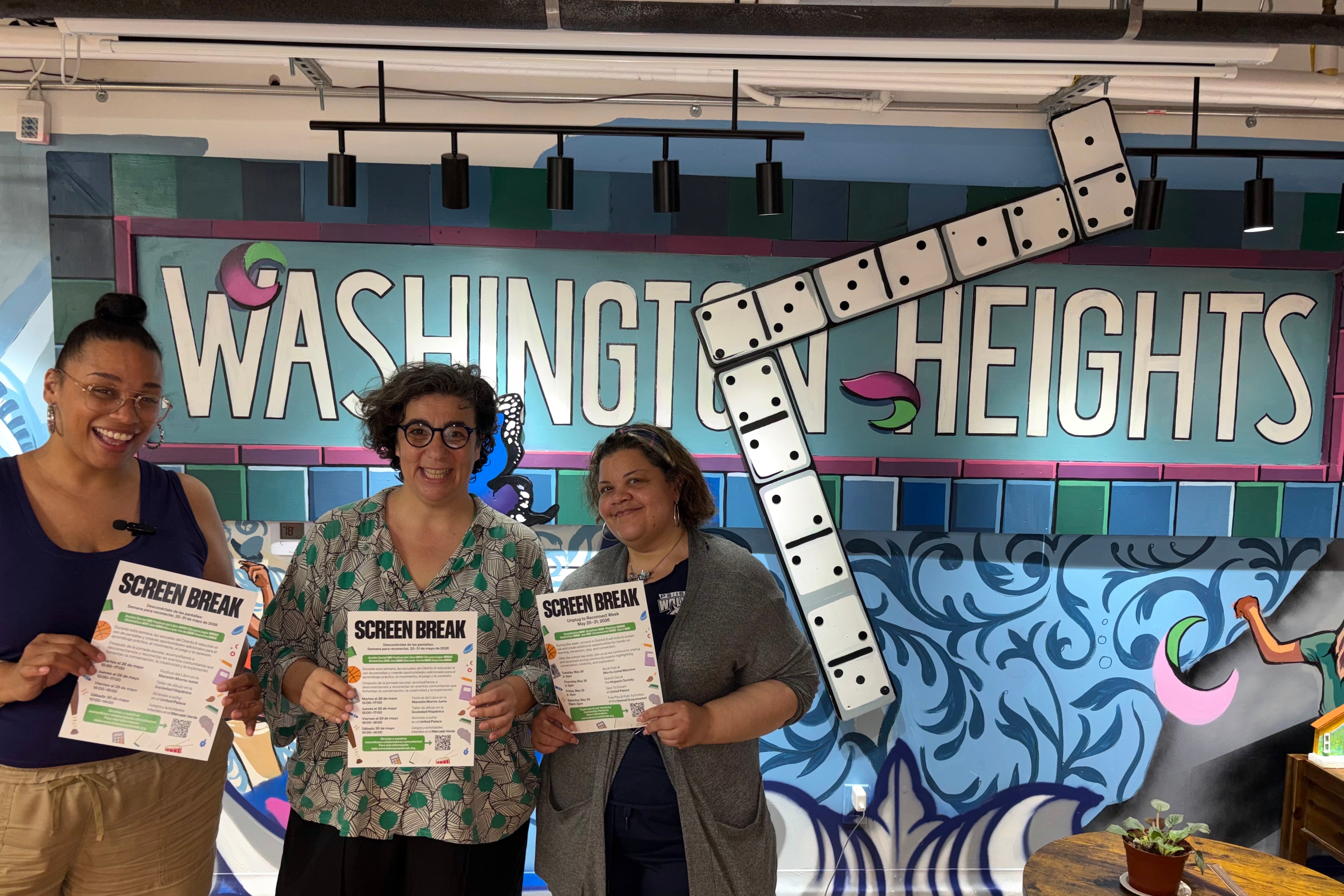 A photograph of three adult women posing for a photograph while holding up a piece of paper in front of a mural that reads "Washington Heights."