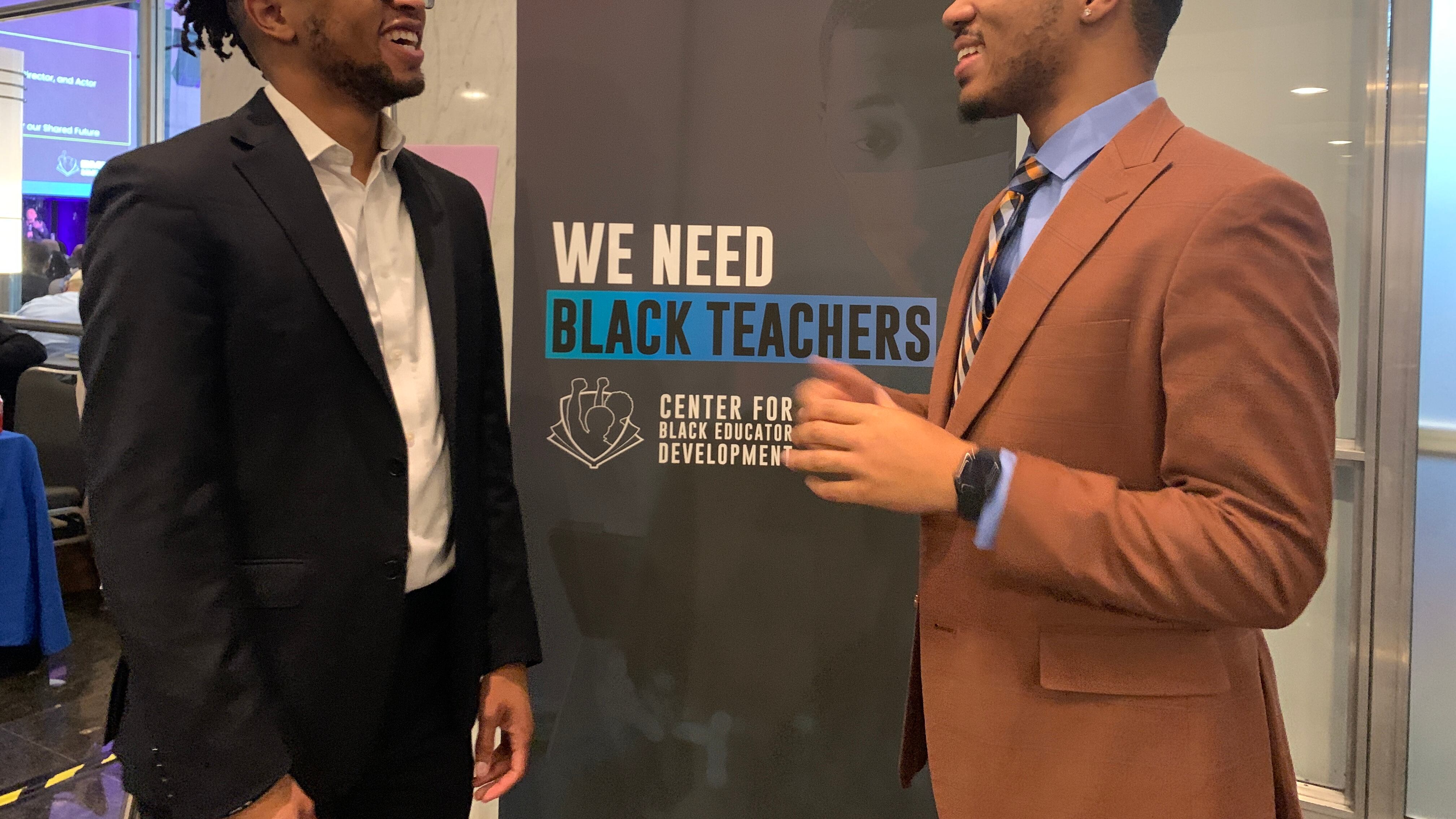 Two Black men - one in a dark jacket and the other in a tan jacket - talk in front of a sign that says “We Need Black Teachers.”