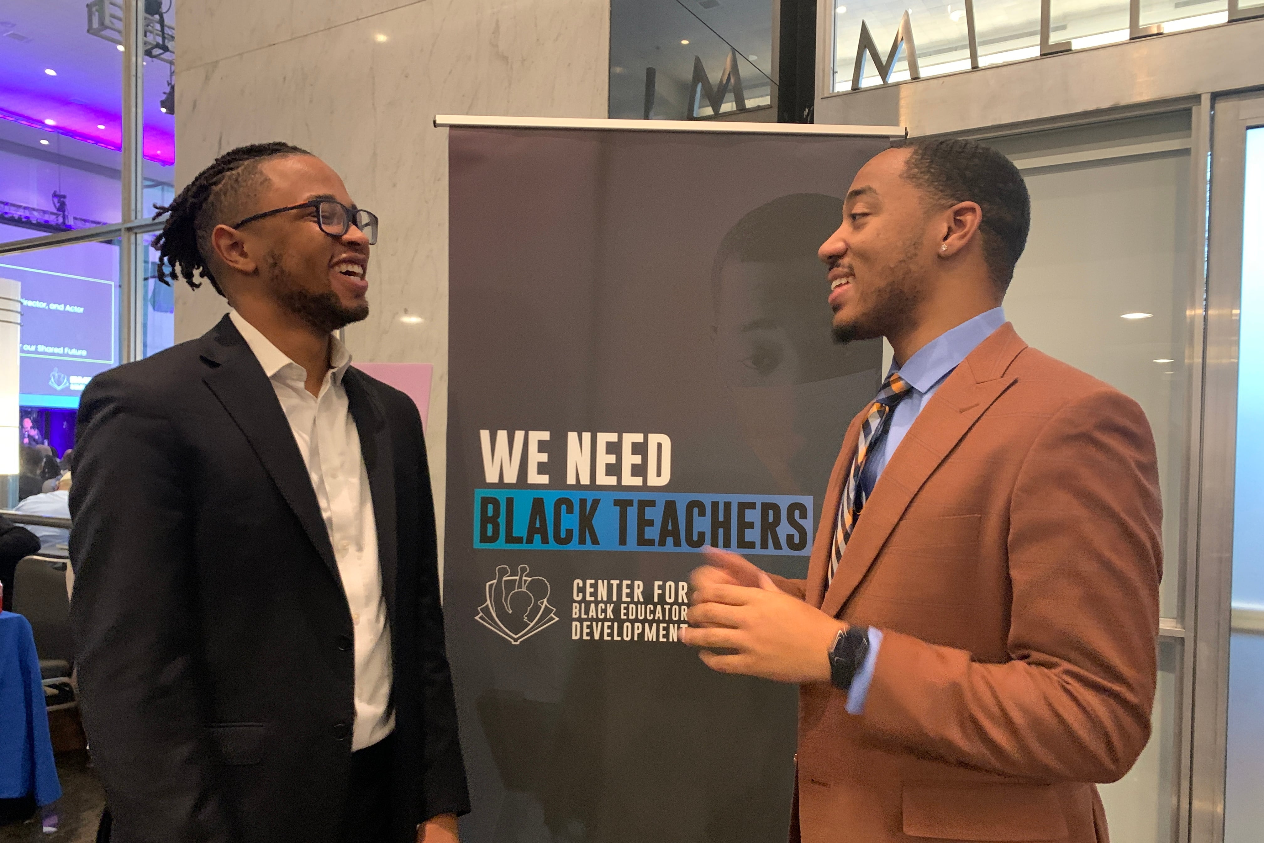 Two Black men - one in a dark jacket and the other in a tan jacket - talk in front of a sign that says “We Need Black Teachers.”