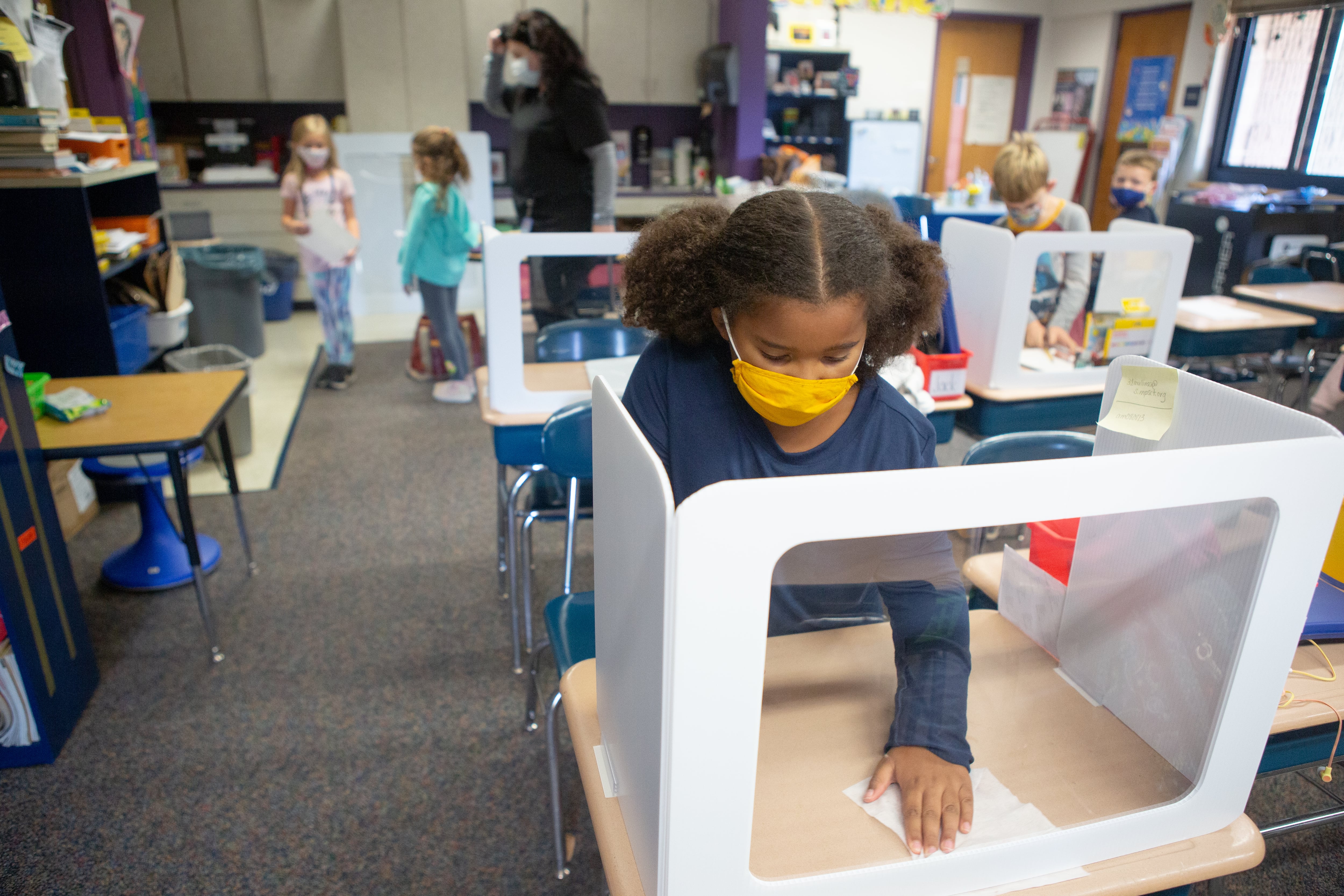 A masked elementary-age student works at a desk with a partition around it, with an adult and two students standing in the aisle in the background, and other students at their desks. All are wearing masks.