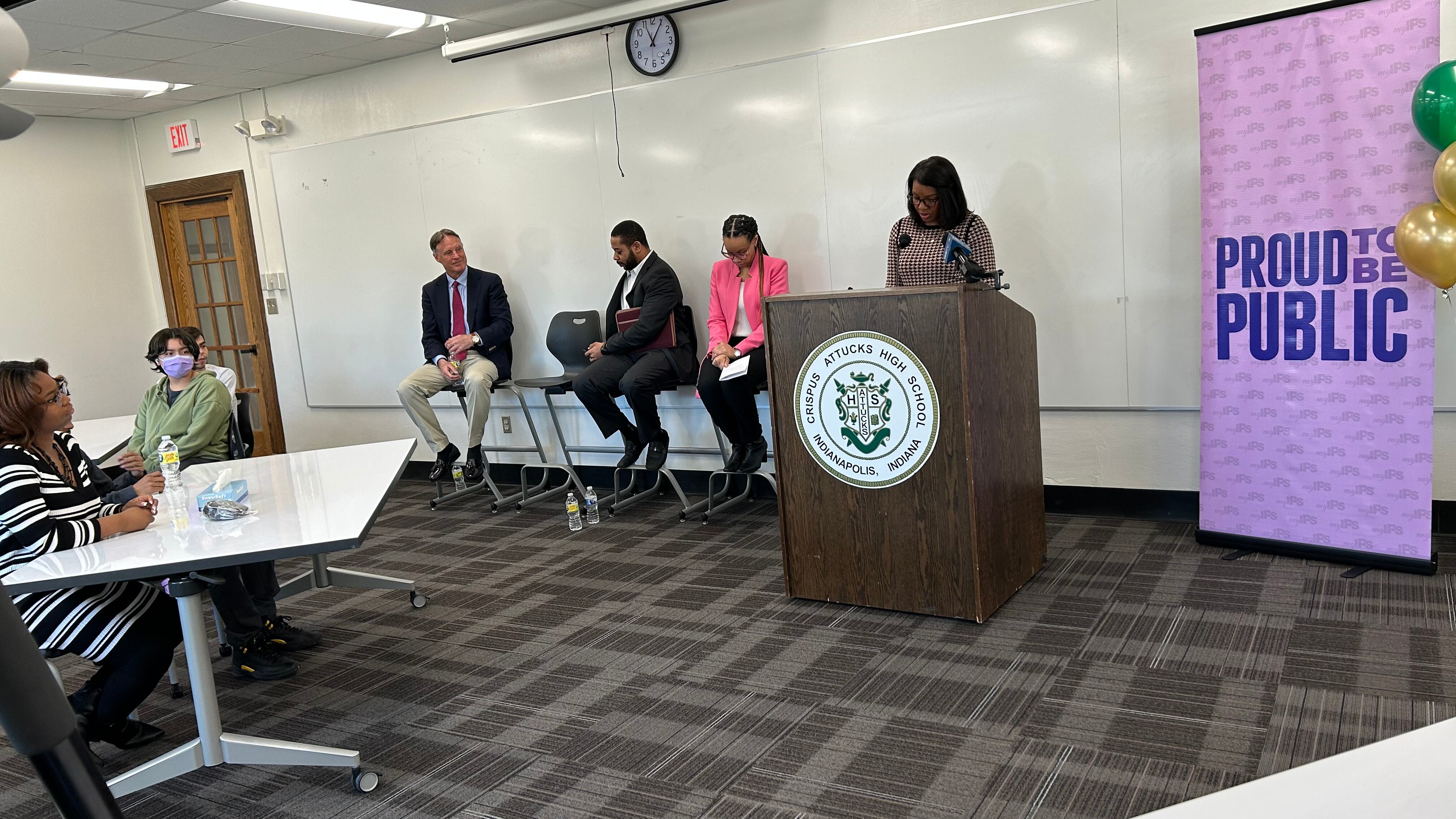 A woman stands speaking behind a podium that reads “Crispus Attucks High School.” On the left are three people sitting down facing an audience. On the right is a banner that says “Proud to be public.”