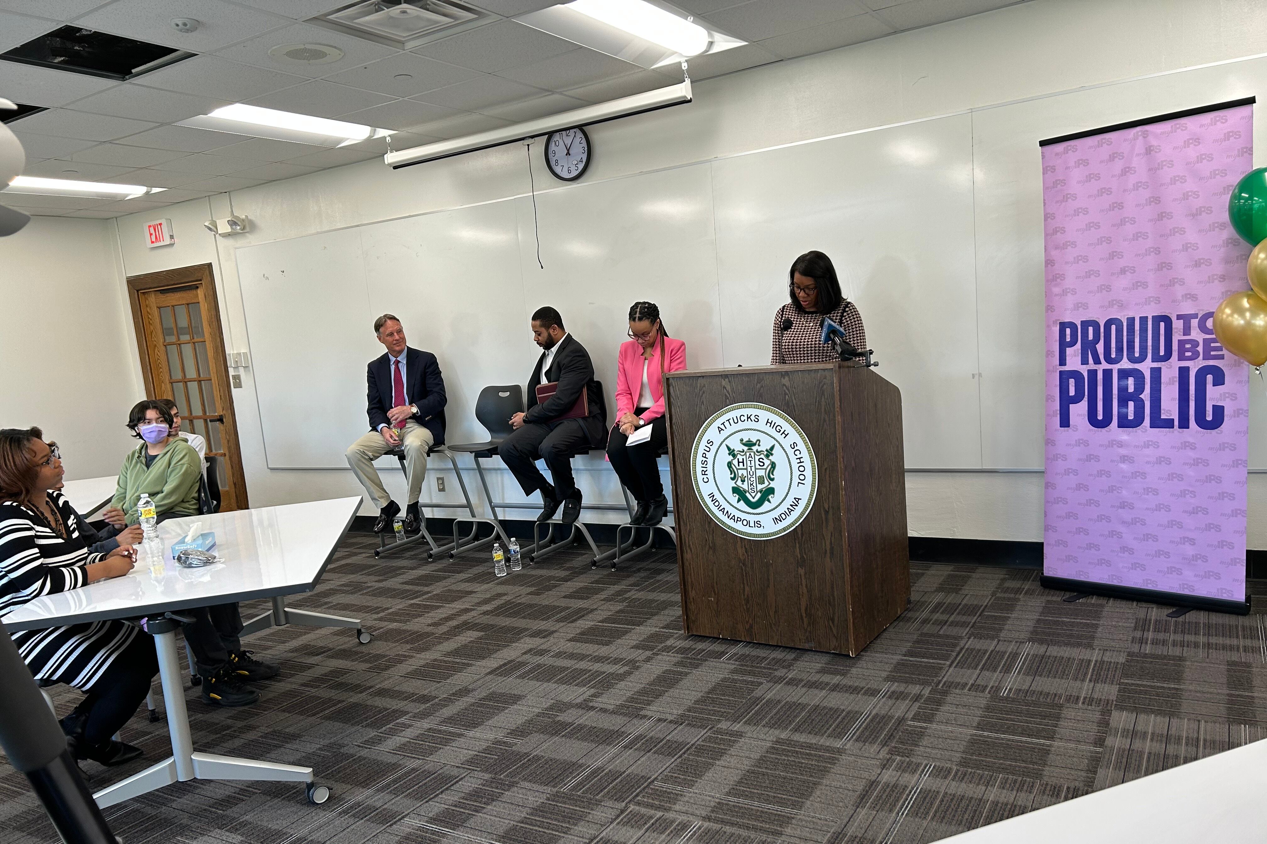 A woman stands speaking behind a podium that reads “Crispus Attucks High School.” On the left are three people sitting down facing an audience. On the right is a banner that says “Proud to be public.”