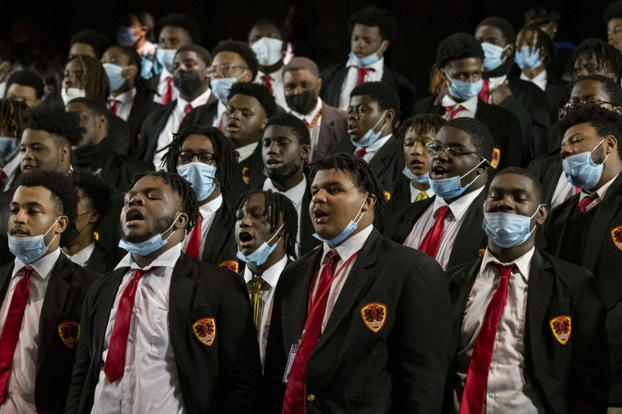 A large group of high school students all wearing the same uniform and have masks around their chins sing in a choir.