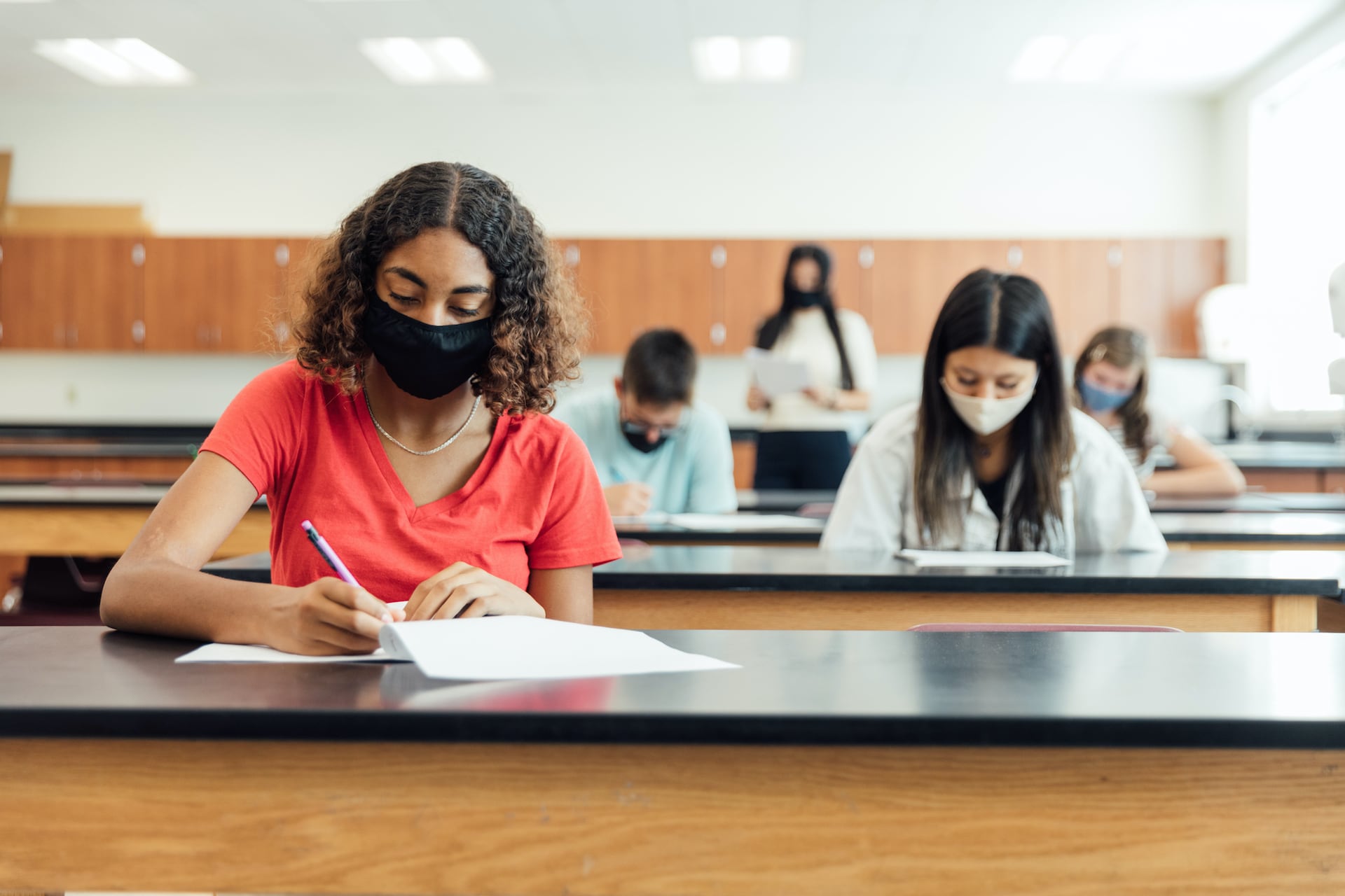 Masked high school students sit several feet apart at long tables working on papers.