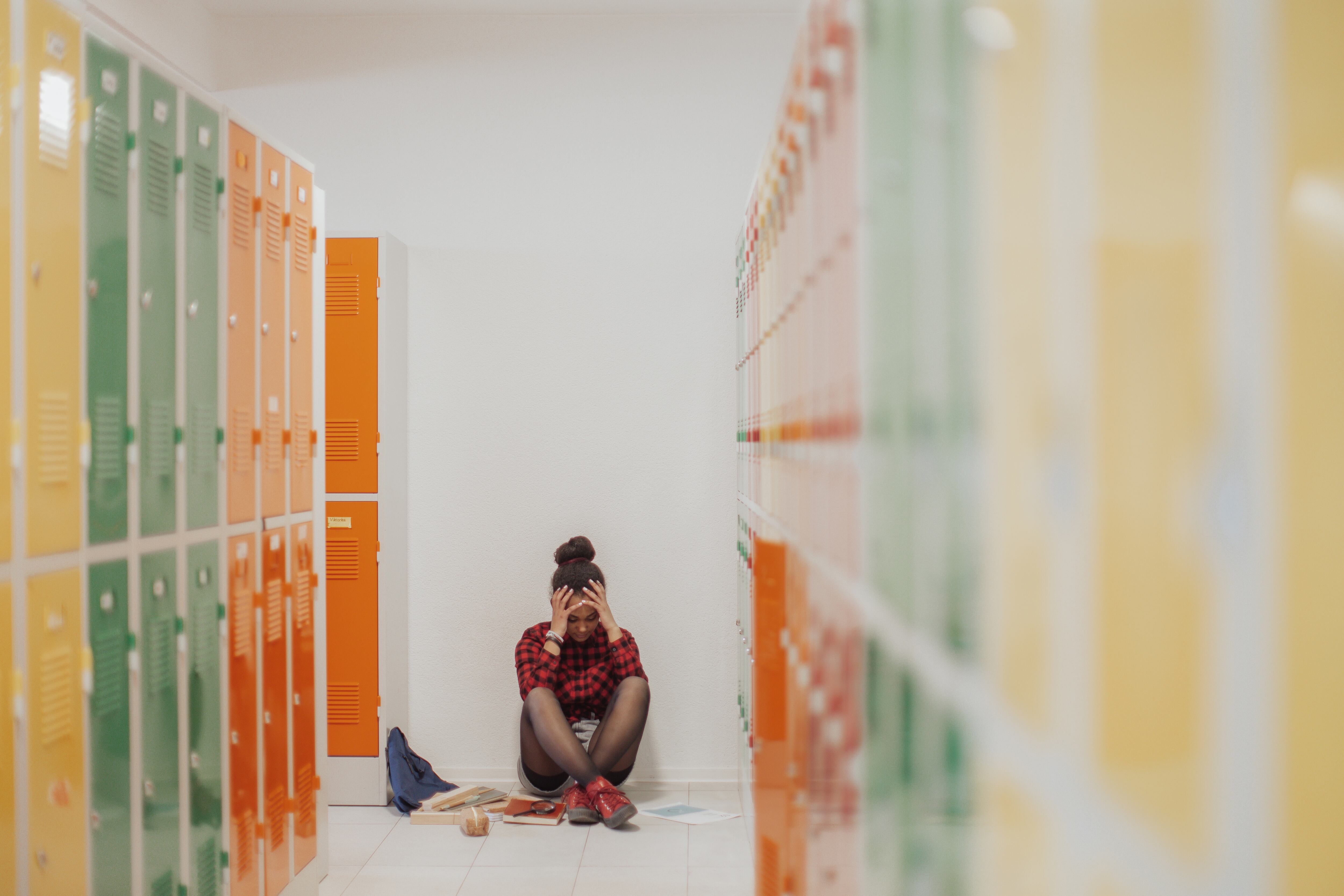 A high school girl sits with her hands covering her face on the floor of a hallway with colorful lockers on both sides.
