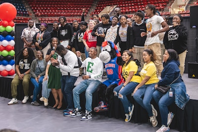 A couple dozen high school students are sitting and standing in an auditorium, facing a camera that is off to the side.