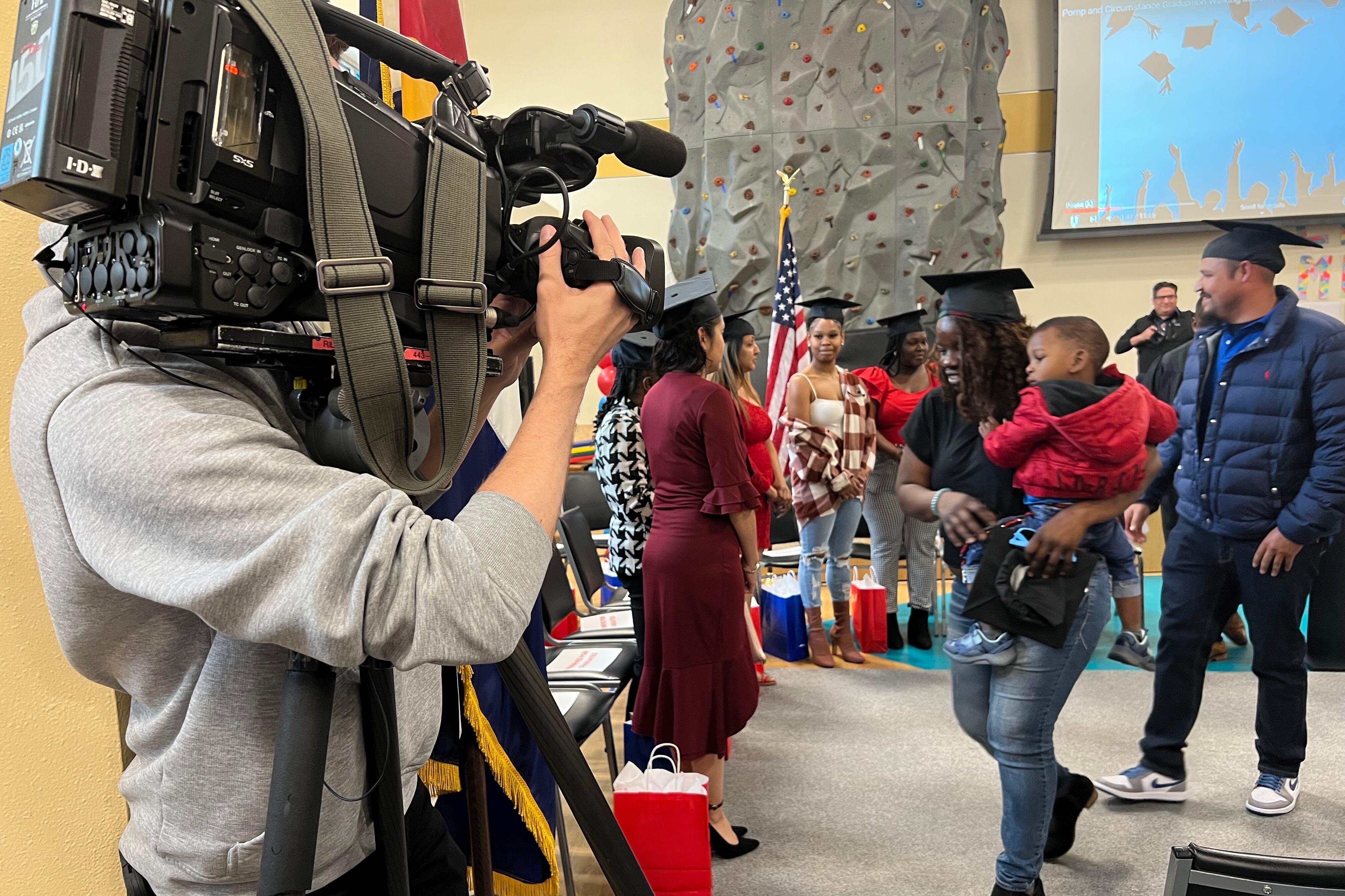Adults wearing black mortarboards walk into a gymnasium. A TV camera captures the scene.