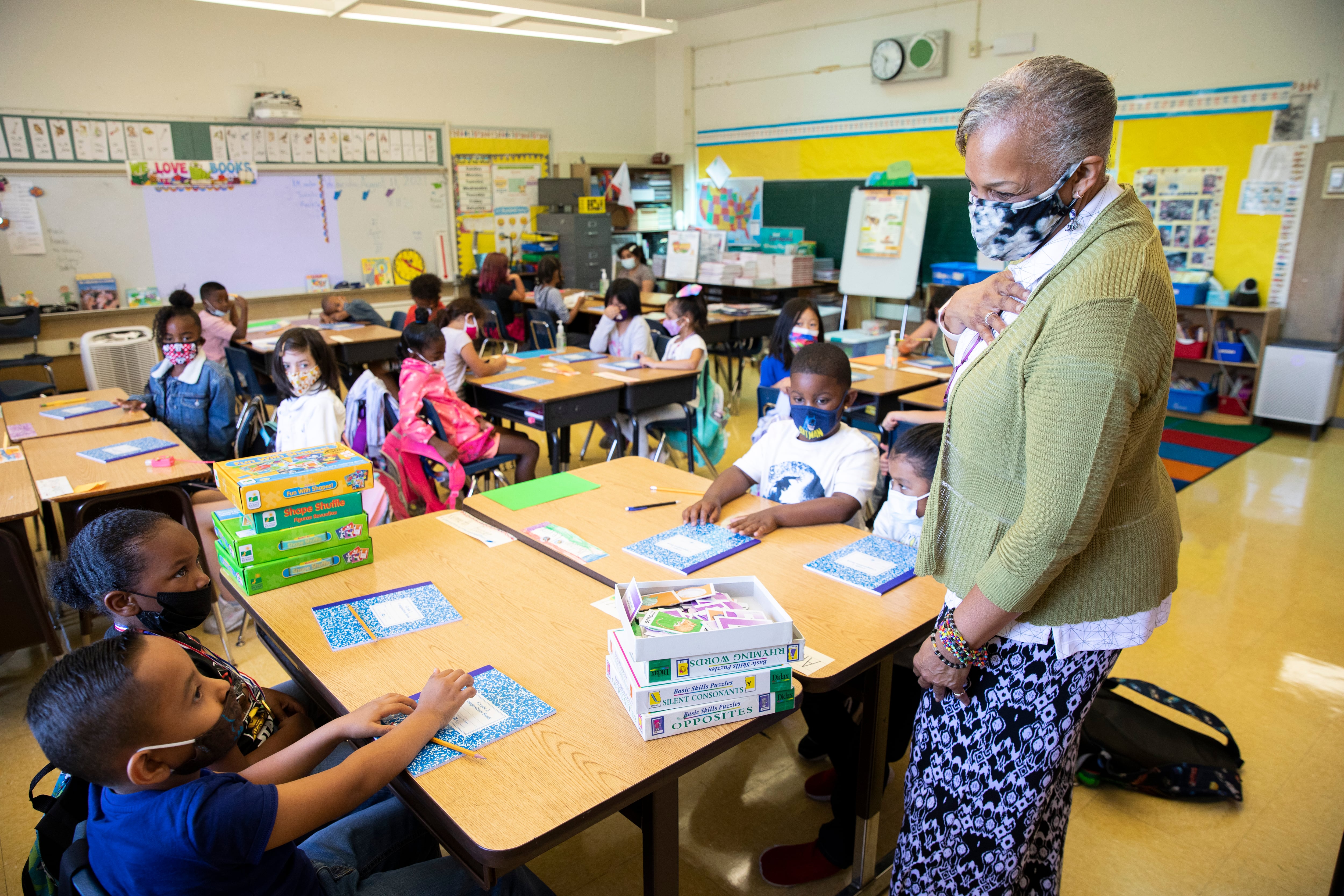 An older female elementary teacher gestures at a table of young students in her full classroom, all of them wearing masks due to COVID protocols.