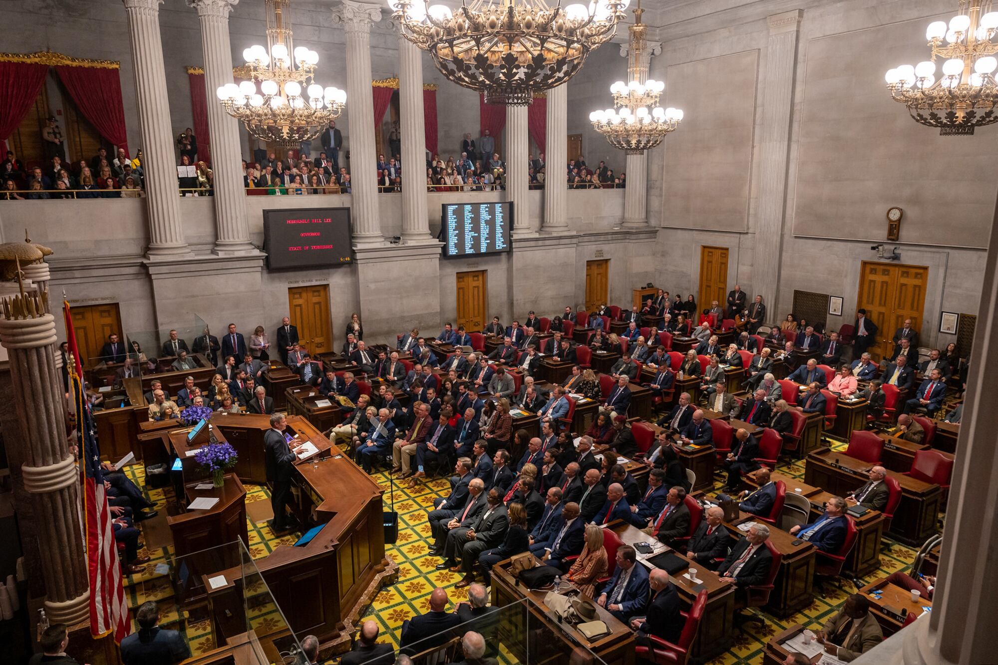 A large room with two levels is filled with dozens of people listening to a person who is standing at a wooden podium on the left.